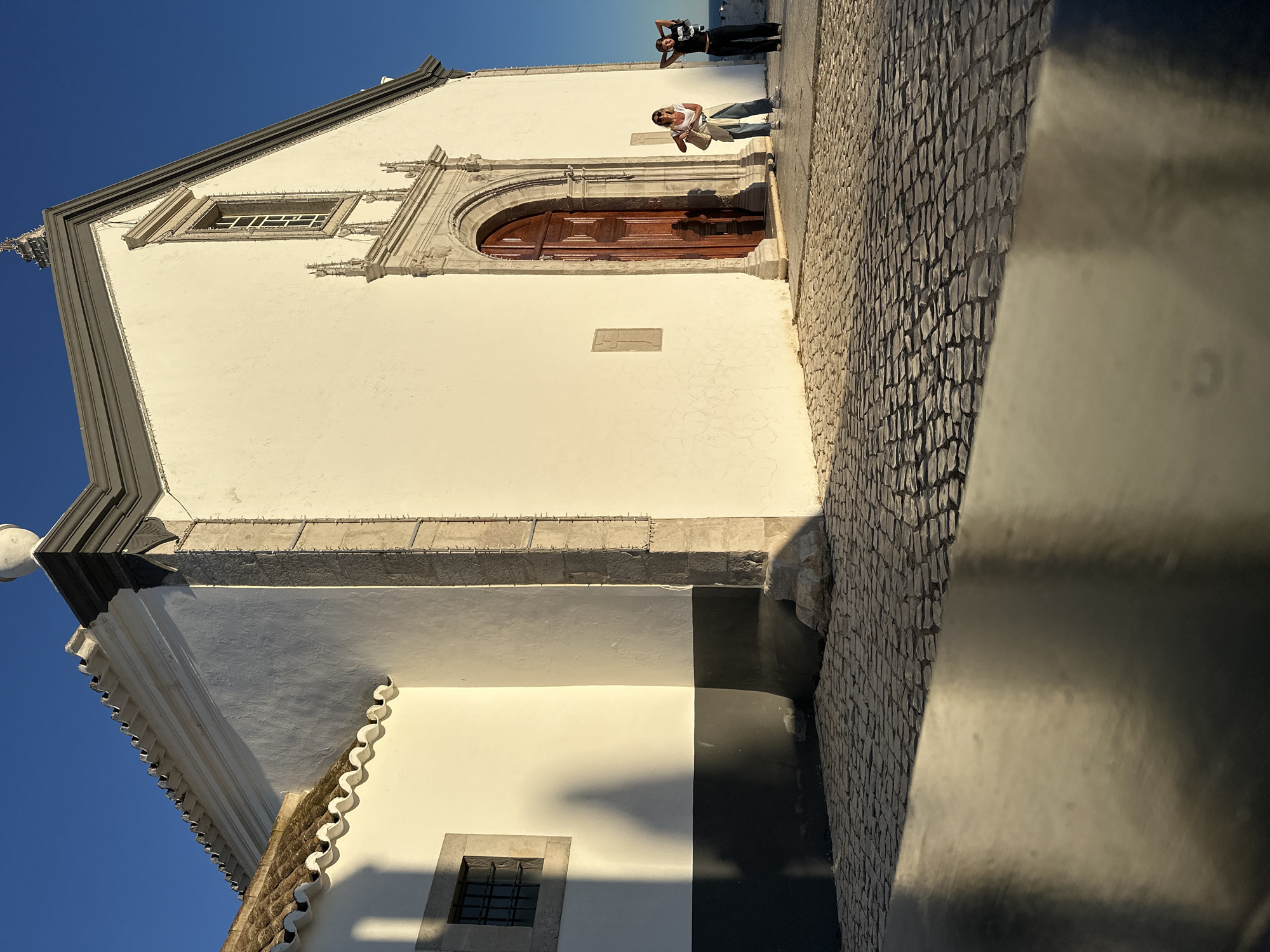 Exterior shot of a church in Vila Real de Santo António, Portugal, taken during daylight hours. The church's facade is predominantly white with a gray roof and stone accents around the windows and a wooden door. Two individuals stand near the door, one appearing to take a picture of the other. A cobblestone pavement extends alongside the building, with a curb in the foreground. The sky is clear and blue.