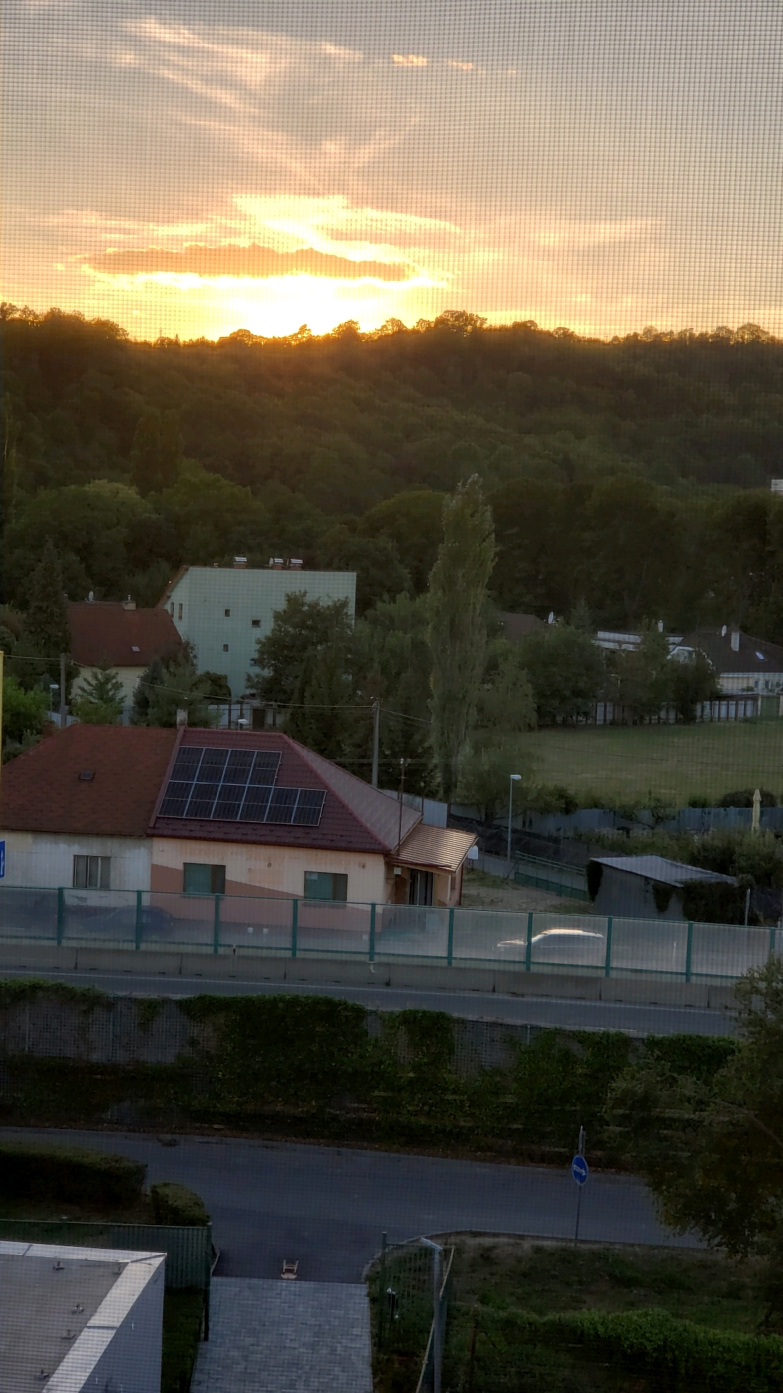 A vertical, eye-level outdoor shot captures a sunset view over a residential area and a wooded hillside, seen through a mesh screen. The sky above is a pale orange and yellow gradient, with wispy clouds illuminated by the setting sun. A dense, dark green forest covers a sloping hillside that dominates the middle ground.
In the foreground, a two-story residential building with a reddish-brown tiled roof is visible. Solar panels are installed on the roof of this building. Adjacent to it, a lighter colored building with a flat roof and a light green facade can be seen. A paved road runs horizontally across the lower middle section of the image, bordered by a green mesh fence. Further down, another road is visible with a blue traffic sign indicating a one-way street.
The scene appears to be in the city of Prešov, Slovakia, based on contextual information not visible in the image itself. There are no people visible in the frame. The overall atmosphere is calm and serene, with the golden light of sunset casting long shadows and highlighting the textures of the landscape and buildings.