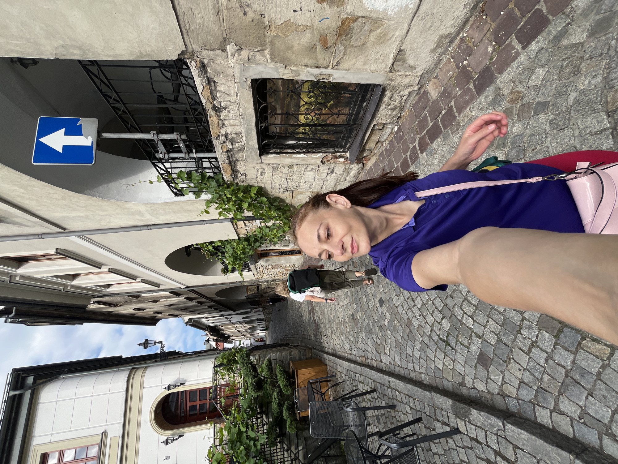 In the city of Unknown, Unknown, a woman with brown hair, wearing a purple polo shirt, red pants, and a pink purse, walks down a cobblestone street. She is holding her phone to take a selfie, her arm outstretched. Behind her, another person wearing a backpack walks away from the camera, while another individual stands further down the street. The street is lined with old buildings, some with arched windows and iron railings. A blue and white traffic sign with an upward-pointing arrow is visible on the side of a building. The environment appears to be an urban setting with a historic feel.