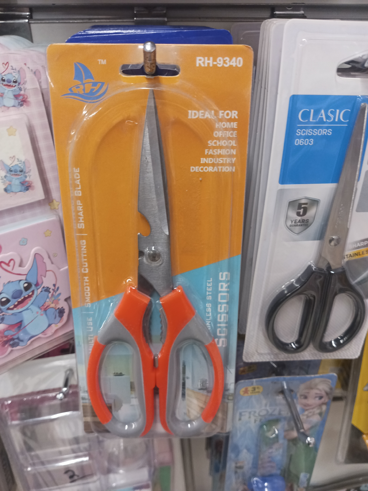 The image shows a close-up, eye-level shot of a retail display of scissors in packaging. The primary focus is on two sets of scissors, one with orange and grey handles, packaged in an orange and blue blister pack with the text "RH-9340" and "IDEAL FOR HOME OFFICE SCHOOL FASHION INDUSTRY DECORATION". The other set of scissors, to the right, has black handles and is packaged in a white and blue blister pack with the brand name "CLASC" and "SCISSORS 0603". There is also a small badge indicating "5 YEARS GUARANTEE". To the left of the main focus, there are other packaged items, including what appears to be stationery with cartoon characters, possibly Stitch from Disney's Lilo & Stitch. The background shows the shelving of a store. The lighting is artificial and appears to be from overhead fluorescent lights. This image does not provide any information about the city of Peshawar City Tehsil, Pakistan.