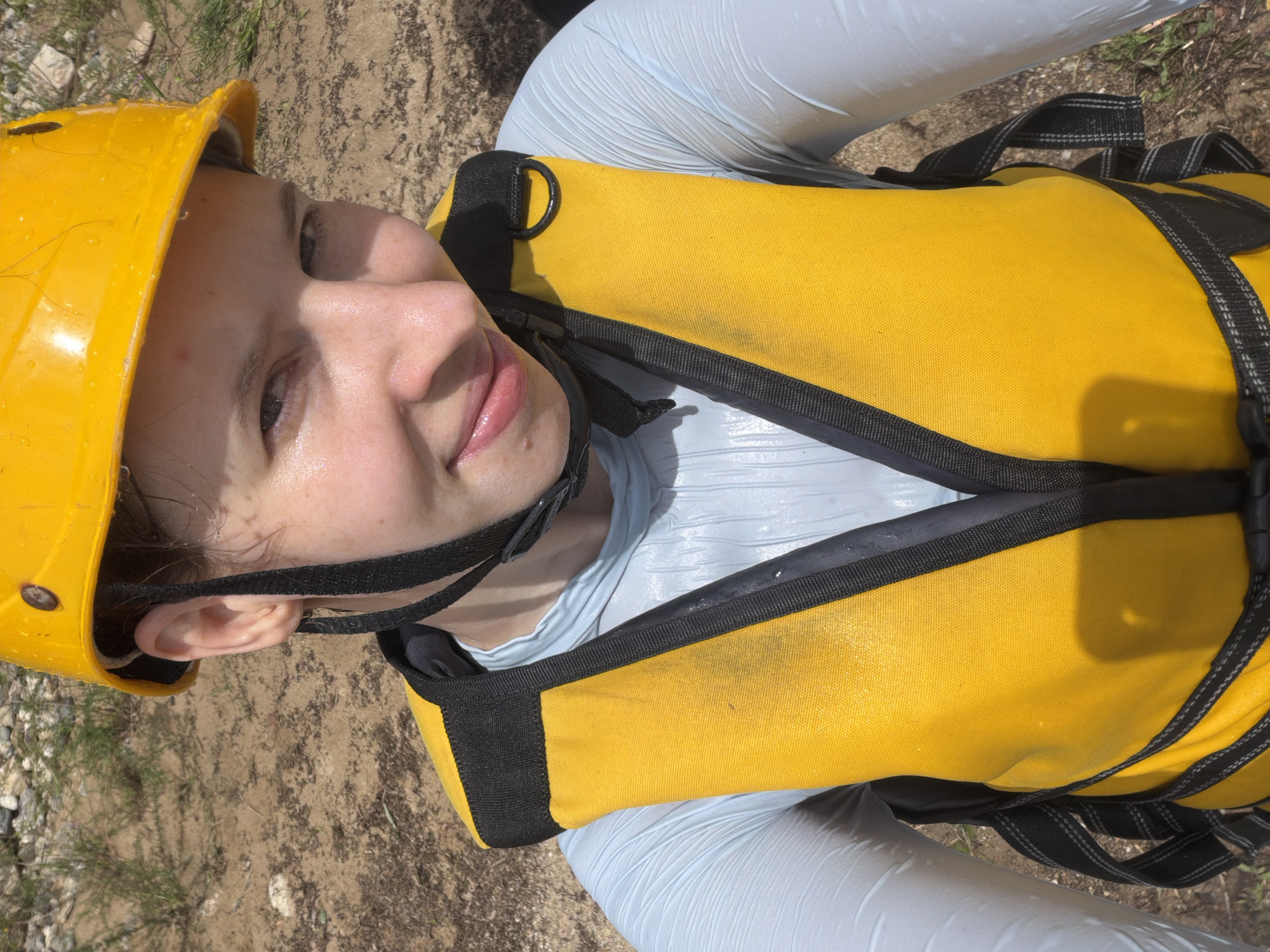 This is a close-up, top-down shot of a young person wearing a yellow safety helmet and a bright yellow life vest. The person is looking upwards, with their face partially obscured by the helmet and the life vest straps. Their skin appears slightly flushed and possibly sweaty, suggesting they might be engaged in an outdoor activity. Their lips are slightly parted, giving a hint of a smile.
The setting appears to be outdoors, on a dirt and gravelly ground with some sparse green vegetation visible. The lighting suggests it is daytime, likely sunny, as there are distinct shadows cast on the life vest. The overall impression is of someone preparing for or participating in an adventure sport, possibly related to water activities given the life vest. The person's gaze and slight smile convey a sense of anticipation or enjoyment. There is no visible text in the image.