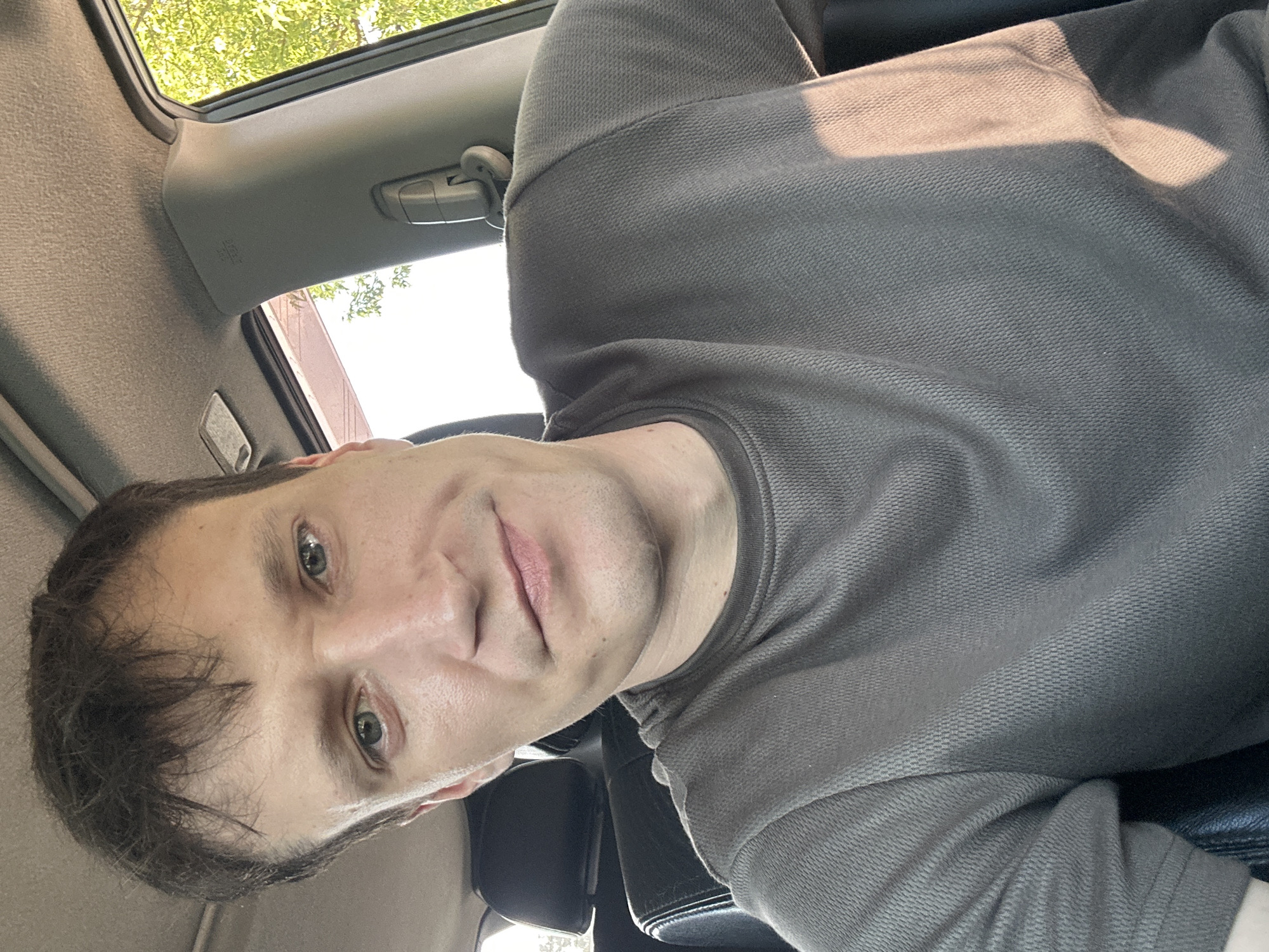 A medium shot, selfie-style photo taken from the passenger seat of a vehicle shows a fair-skinned man with short, dark brown hair and blue-green eyes. He is looking directly at the camera with a neutral expression. He is wearing a dark grey, textured t-shirt. The interior of the vehicle is visible, including the headrest of the seat behind him, the car's headliner and pillar, and a glimpse of the outdoor environment through the window. The lighting suggests it is daytime, possibly late afternoon due to the warm sunlight casting shadows on his shirt and the vehicle's interior. The man appears to be in Makiv, Ukraine, though no specific landmarks are identifiable to confirm the exact location within the city.