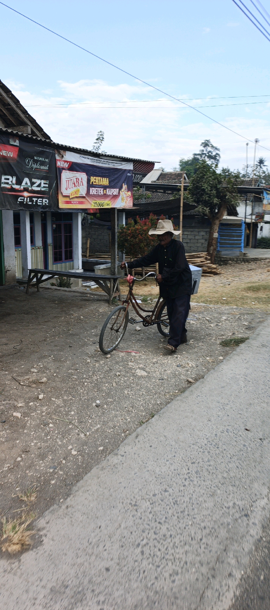 In Kasiyan, Indonesia, an elderly man wearing a wide-brimmed hat and dark clothing is standing next to a bicycle. The bicycle, which appears to be an older model with a brown frame and a metal carrier attached to the back, is leaning on its kickstand. The man is holding the handlebars, and his right foot is on the ground.
The scene is set in front of a building with a roof extending outwards, under which several banners are displayed. One banner is black with red and white lettering that reads "BLAZE FILTER". Another banner, predominantly red and white, features text that includes "JUARA PERTAMA KRETEK KAPSUL" and a price of "17.000". To the right of the building, there are piles of lumber stacked outdoors.
The ground is a mixture of gravel and dirt, with a paved road running diagonally across the foreground. In the background, other buildings and trees are visible, under a sky with scattered clouds. A wooden bench is positioned in front of the building. The overall impression is a rural or semi-urban setting in Indonesia.
