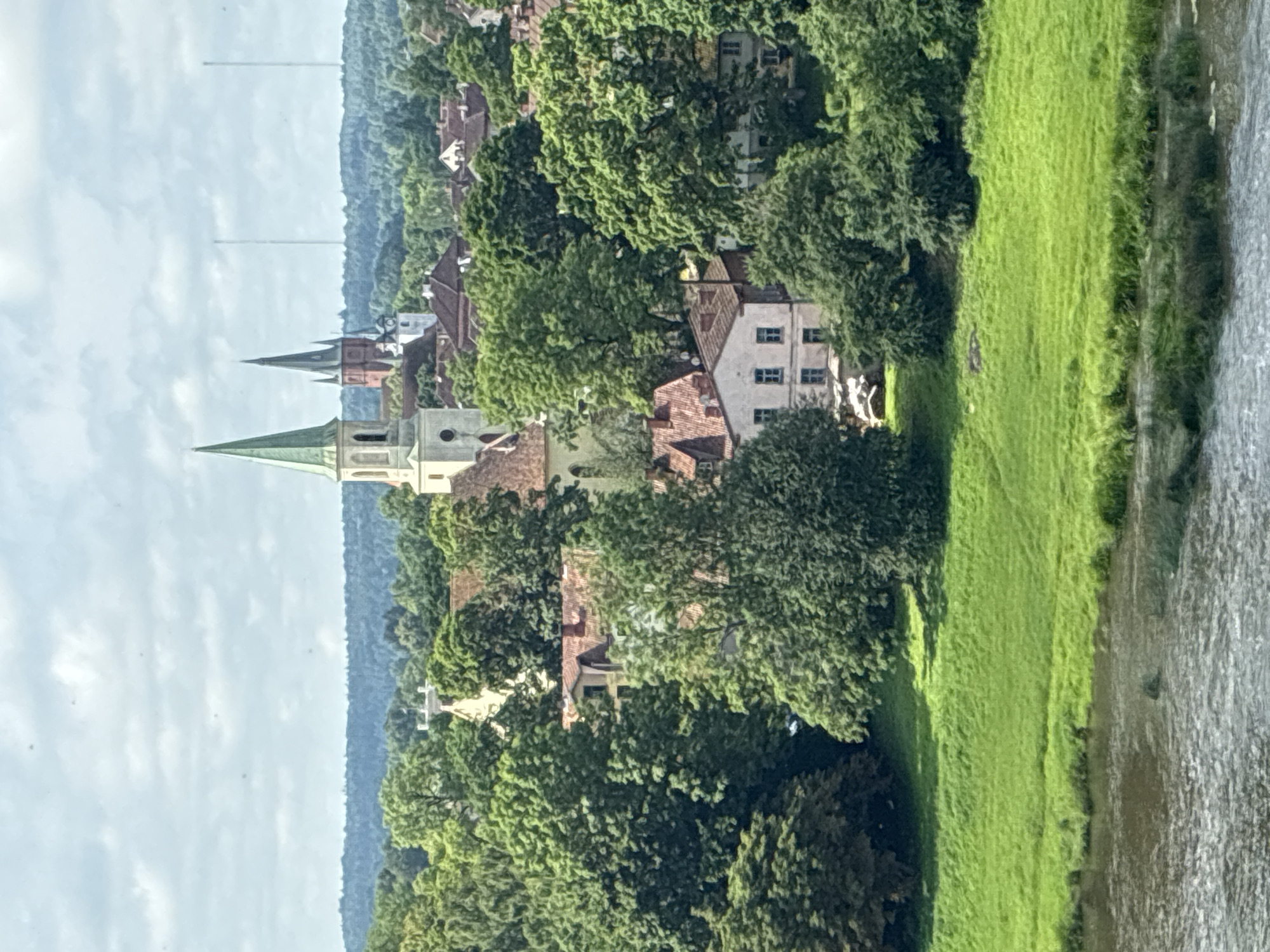 The image shows a panorama of Kuldīga, Latvia, with a focus on the historic town center. A wide river flows in the foreground, bordered by a lush green embankment. Beyond the embankment, the town rises, characterized by a dense canopy of mature trees interspersed with red-tiled roofs and various buildings. Prominent church spires, notably a tall, pale spire with a green roof and a darker, gothic-style spire behind it, punctuate the skyline, indicating the presence of religious structures. Residential and municipal buildings, with cream-colored facades and visible window details, are nestled amongst the foliage. The overall impression is that of a tranquil, verdant European town, likely captured from an elevated vantage point.