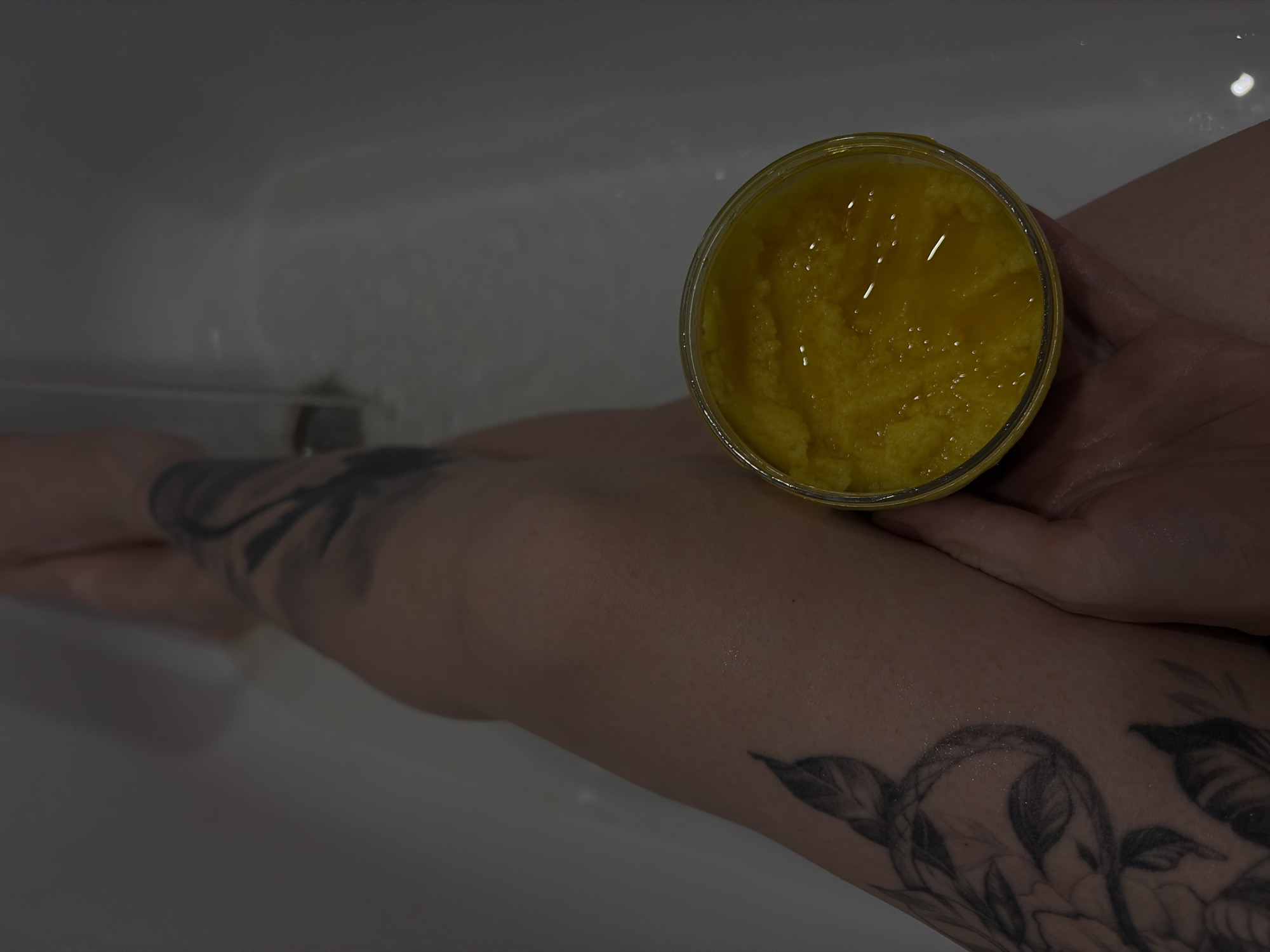 In a bathtub setting, likely indoors and not specifically identifiable to Chaikovsky, Russia, an individual is holding a clear jar filled with a yellow, gel-like substance. The person's arm, which is tattooed with floral designs, is partially submerged in the water of the bathtub. The tub itself is white, and water is visible near the tap. The focus is on the jar of yellow substance and the tattooed arm.