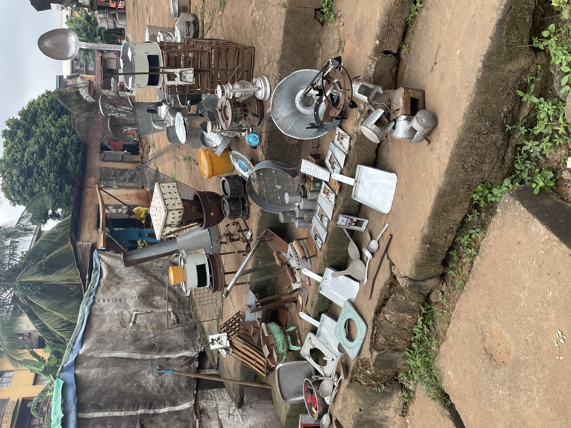 This image depicts an outdoor market scene in Nkpor, Nigeria, likely a section specializing in metal cookware and household items. The foreground is dominated by a collection of various metal objects arranged on concrete steps and the ground. These include numerous pots and pans of different sizes, basins, ladles, spatulas, and what appear to be various types of cooking stoves, some of which are made of metal. There are also items like kerosene lamps, a large spoon, a fan, and a rake. The background shows a rustic, open-air stall with a tarpaulin roof, behind which are buildings and vegetation, including a prominent palm tree. A few individuals are visible in the background, appearing to be customers or vendors, interacting within the market environment. The overall impression is one of a bustling, informal marketplace offering a variety of domestic goods.
