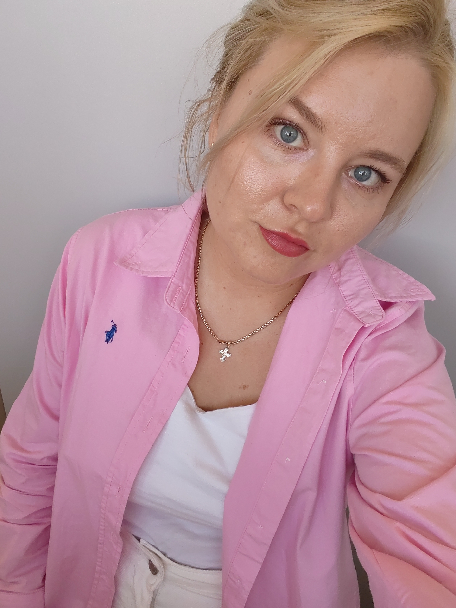 A woman with blonde hair and blue eyes is posing for a selfie. She is wearing a pink collared shirt over a white top and a gold necklace with a cross pendant. The background is a plain white wall. There is no indication of a location in Izhevsk, Russia.