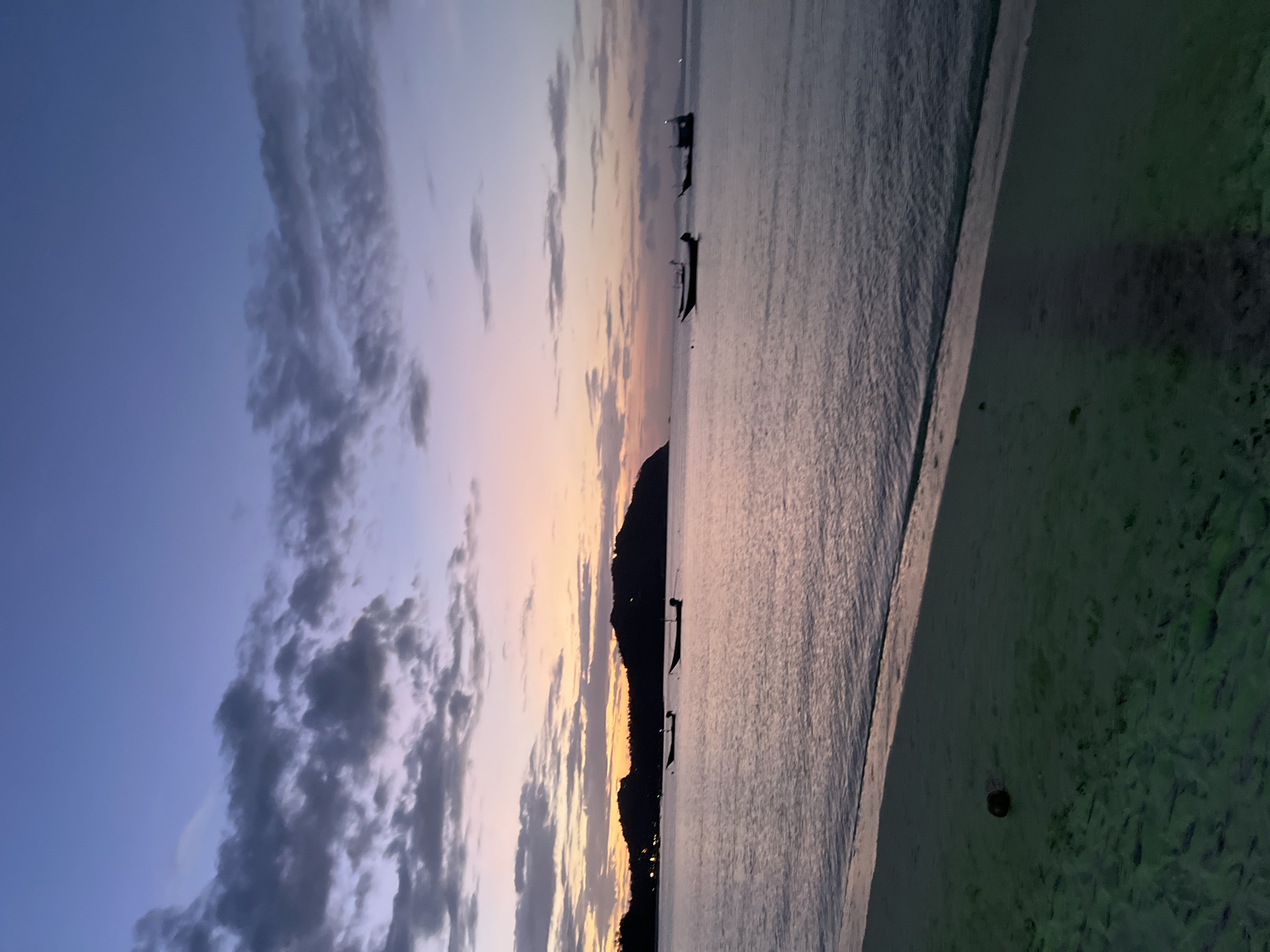 The image depicts a tranquil scene of a beach in the Ko Pha-ngan District of Thailand during twilight. In the foreground, a sandy beach, which appears somewhat damp and textured with footprints, slopes down towards the water. A dark, rounded object, possibly a coconut, rests on the sand.
The sea stretches out to the horizon, its surface reflecting the soft hues of the sky. Gentle ripples indicate a calm body of water. Several small boats are visible on the water, some appearing as dark silhouettes. In the middle ground, a landmass, likely an island or peninsula, rises from the sea, its outline also silhouetted against the sky. Lights are faintly visible along the coastline of this landmass, suggesting a settlement.
The sky is a gradient of colors, transitioning from a deep blue at the top to lighter shades of purple, pink, and orange closer to the horizon. Clouds are scattered across the sky, some appearing dark and textured, while others are lighter and more wispy. The overall atmosphere is serene and peaceful, capturing the transition from day to night over a tropical coastline.