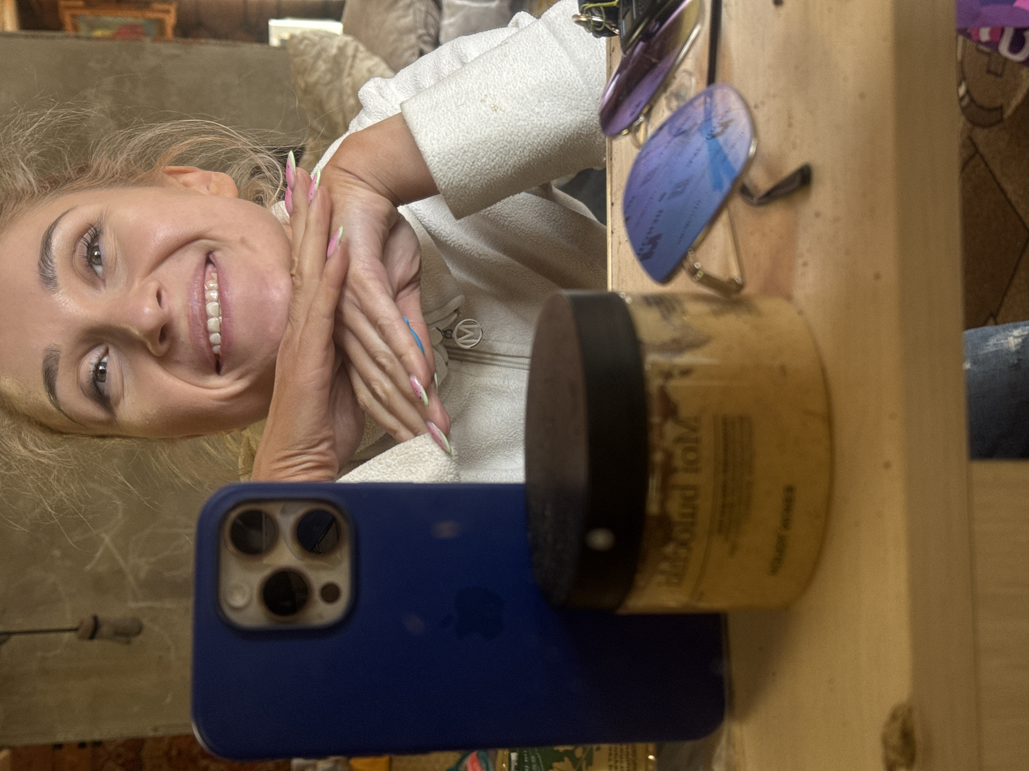 A woman with blonde hair and long, manicured nails is smiling at the camera. Her hands are clasped together in front of her face. She is wearing a white fleece jacket.
A blue iPhone is visible, resting on a wooden surface. A jar with a black lid is near the phone. A pair of sunglasses sits on the surface next to the jar. The image is rotated.
There is no indication of location in the city of Rosstani, Russia.