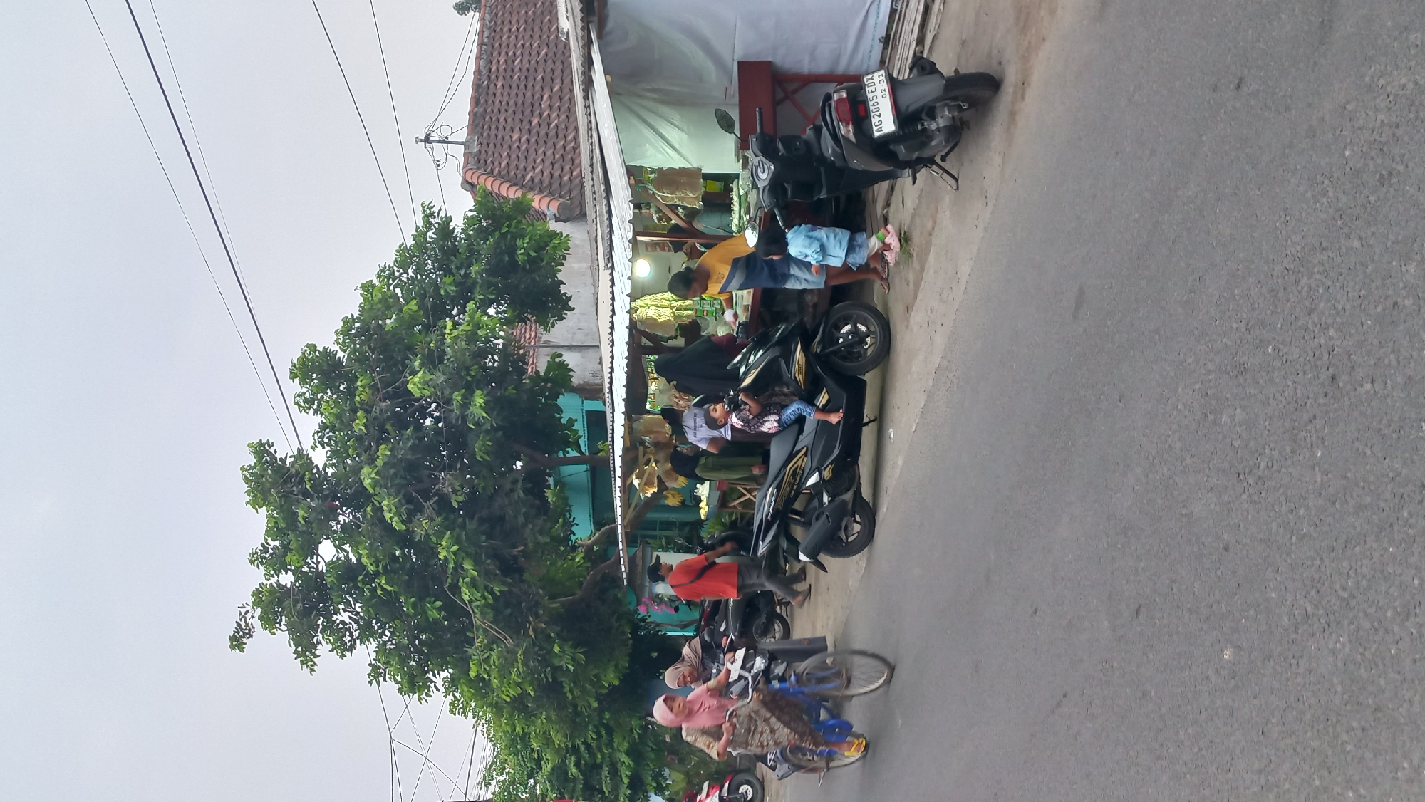 The image captures a street scene in Pare, Indonesia, depicting a modest roadside market. A woman in a pink hijab is riding a blue bicycle on the left side of the paved road. She is wearing a patterned dress and yellow shoes. Behind her, a man in a red shirt walks away from the camera. A black motorcycle is parked on the side of the road, in front of a stall. On the right side of the road, a food stall is set up with a white awning. Several people are gathered around the stall, including a woman in a blue and yellow shirt, and two children. One child, wearing a patterned shirt and blue jeans, is seated on a black motorcycle parked in front of the stall. Another child in a light blue outfit stands near the stall. Merchandise, possibly food items like bananas and dried goods, is displayed on the stall. The environment appears to be an urban or semi-urban area with buildings and trees visible in the background. The sky is overcast.