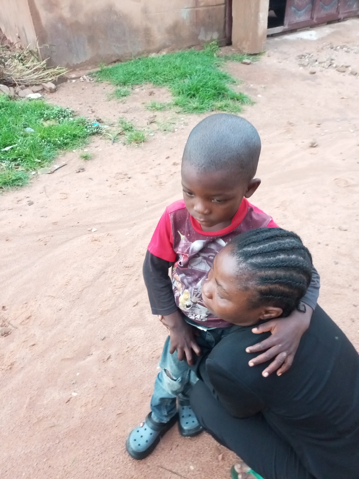 In Tempure, Nigeria, an intimate moment unfolds between a woman and a young boy. The boy, dressed in a red t-shirt with a graphic print and distressed denim jeans, is being embraced by the woman. She is wearing a black outfit and has her hair styled in braids. Their faces are close, suggesting a tender connection, with the boy looking down and the woman looking up at him. The scene appears to be outdoors, with a sandy ground and sparse green grass in the background. A rough, plastered wall and a doorway with a metal gate are visible in the distance, indicating a residential setting. The lighting suggests it could be daytime, possibly late afternoon, with soft shadows. The overall mood is one of warmth and affection.