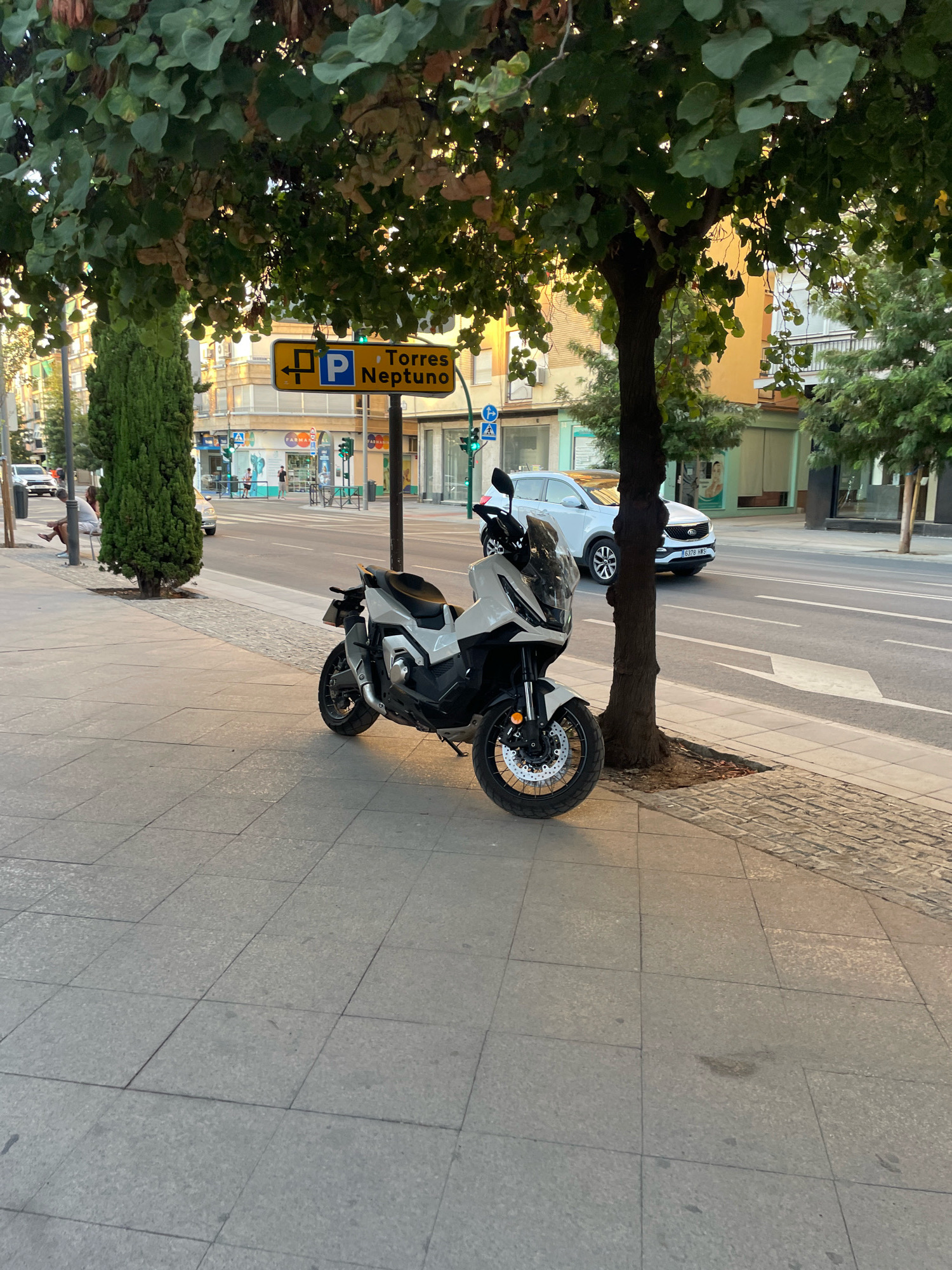 The image depicts a street scene in the city of Granada, Spain, captured from a sidewalk. A white scooter is parked on the paved sidewalk, which is bordered by a street with traffic. In the background, buildings line the street, and trees with green foliage provide shade. A yellow street sign with a blue "P" symbol and the text "Torres Neptuno" is visible, indicating a parking area. A white SUV is driving on the street. The lighting suggests it is either early morning or late afternoon.