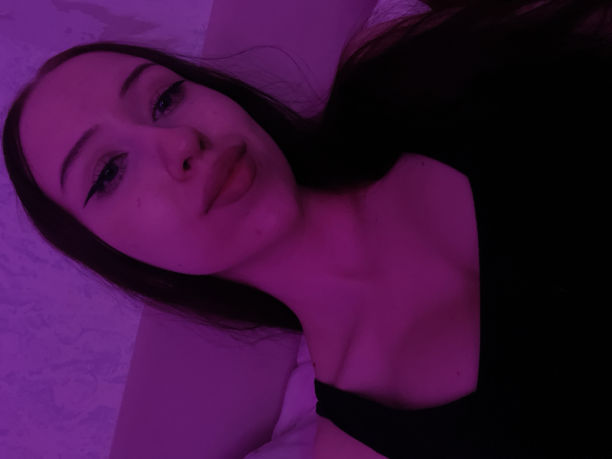 The image is a close-up, head and shoulder selfie of a young woman with long, dark hair. She is wearing a black tank top. Her eyes are open, and she is looking towards the camera. She has winged eyeliner. The lighting in the image is a strong purple hue, suggesting it is either from a colored light source or post-processing. The background appears to be a textured wall, also illuminated by the purple light. There are no identifiable landmarks or elements that would suggest a specific location within Votkinsk, Russia.