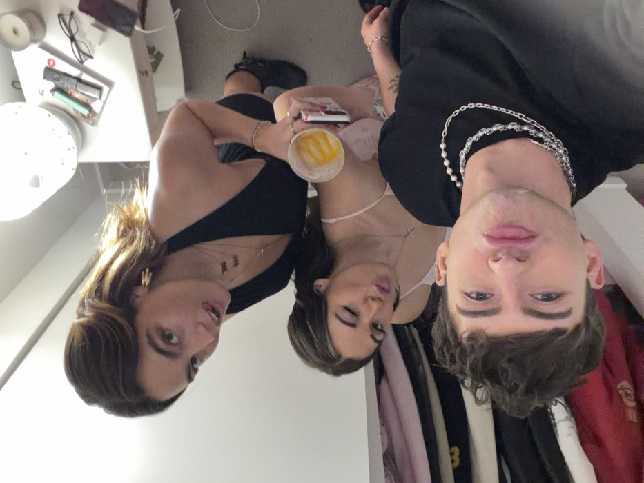 A photo shows three people posing closely together. A woman with brown hair and a black top is on the left. In the center, a woman with brown hair and a light pink top winks while holding a drink and a phone. On the right is a man with dark hair wearing a black shirt and multiple silver necklaces. They are indoors with a cluttered background that includes clothing and a bedside table.