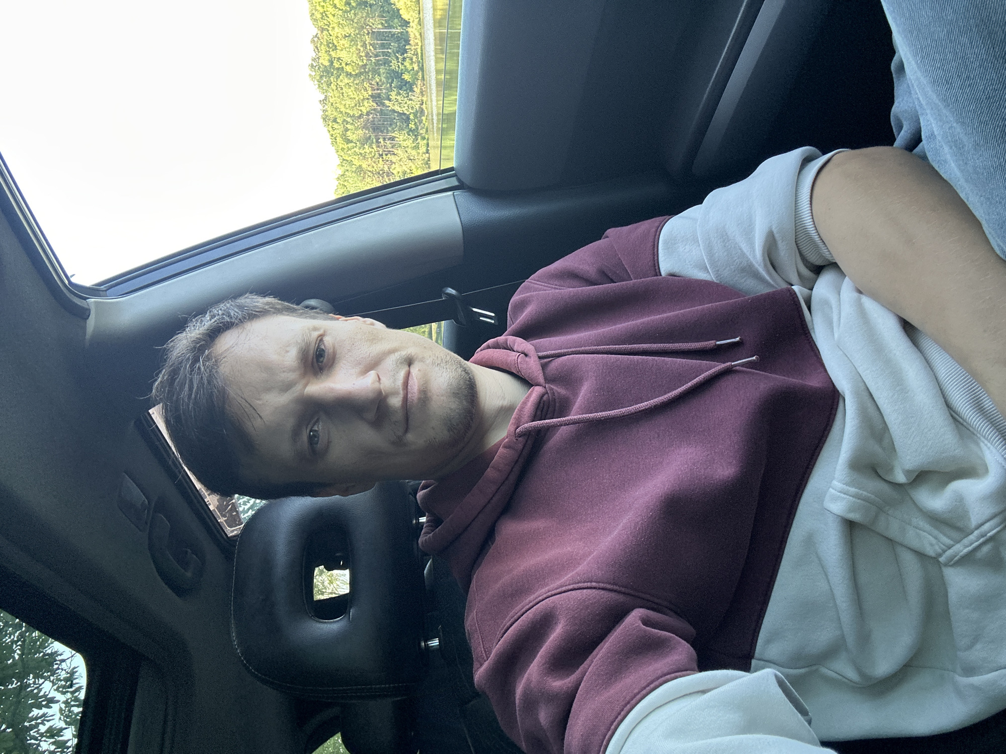 A man is sitting in the driver's seat of a car, looking at the camera. The car's sunroof is open, and sunlight is coming through. Outside the car, there is a body of water and trees. The man is wearing a maroon and cream hoodie and blue jeans. The car's interior is visible, including the headrest, dashboard, and door panel. The scene appears to be set in a rural or natural environment, possibly on the outskirts of Makiv, Ukraine.
