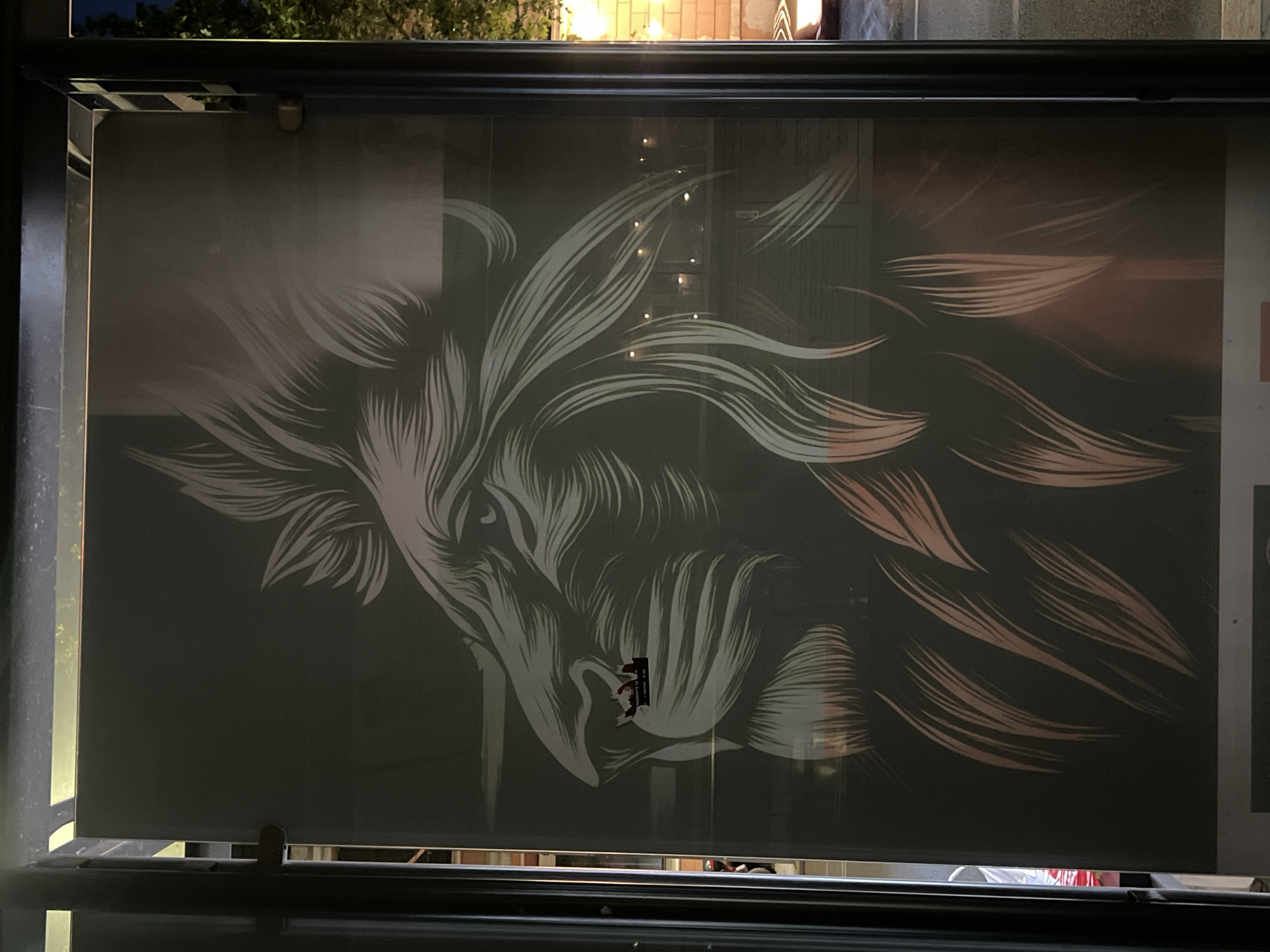 The image shows a large poster or graphic applied to a window, visible from the outside. The artwork depicts a stylized profile of a lion's head, with flowing mane and intricate linework. The mane uses a combination of white and reddish-brown lines to create a sense of depth and movement. A small rectangular sticker or patch is visible on the lion's face near the mouth. The window is framed by a black metal structure with horizontal bars. The ambient light suggests it is nighttime or dusk. The location cannot be determined.