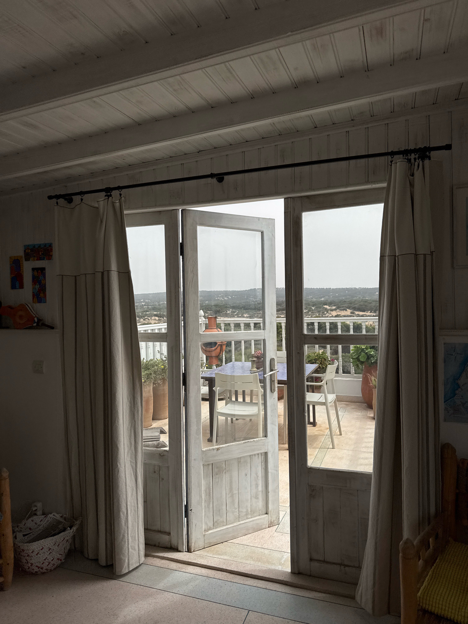 The image depicts an interior view looking out through open double doors onto a balcony and a landscape beyond, likely in Essaouira, Morocco. The interior features a white slatted ceiling and walls, with a dark curtain rod and beige curtains hanging to the sides of the doorway. On the left interior wall, there are three small, colorful abstract artworks. Near the doorway, on the floor, is a woven basket containing what appears to be rolled-up papers.
The balcony, viewed through the open doors, has a light-colored tiled floor and a white railing. On the balcony, there is a blue-tiled table with two white chairs, and a large terracotta pot containing a green plant. Beyond the balcony, a hazy landscape is visible, characterized by undulating terrain covered with sparse vegetation and trees, suggesting a rural or semi-rural setting. The sky is overcast. The overall impression is one of a relaxed, indoor-outdoor living space.