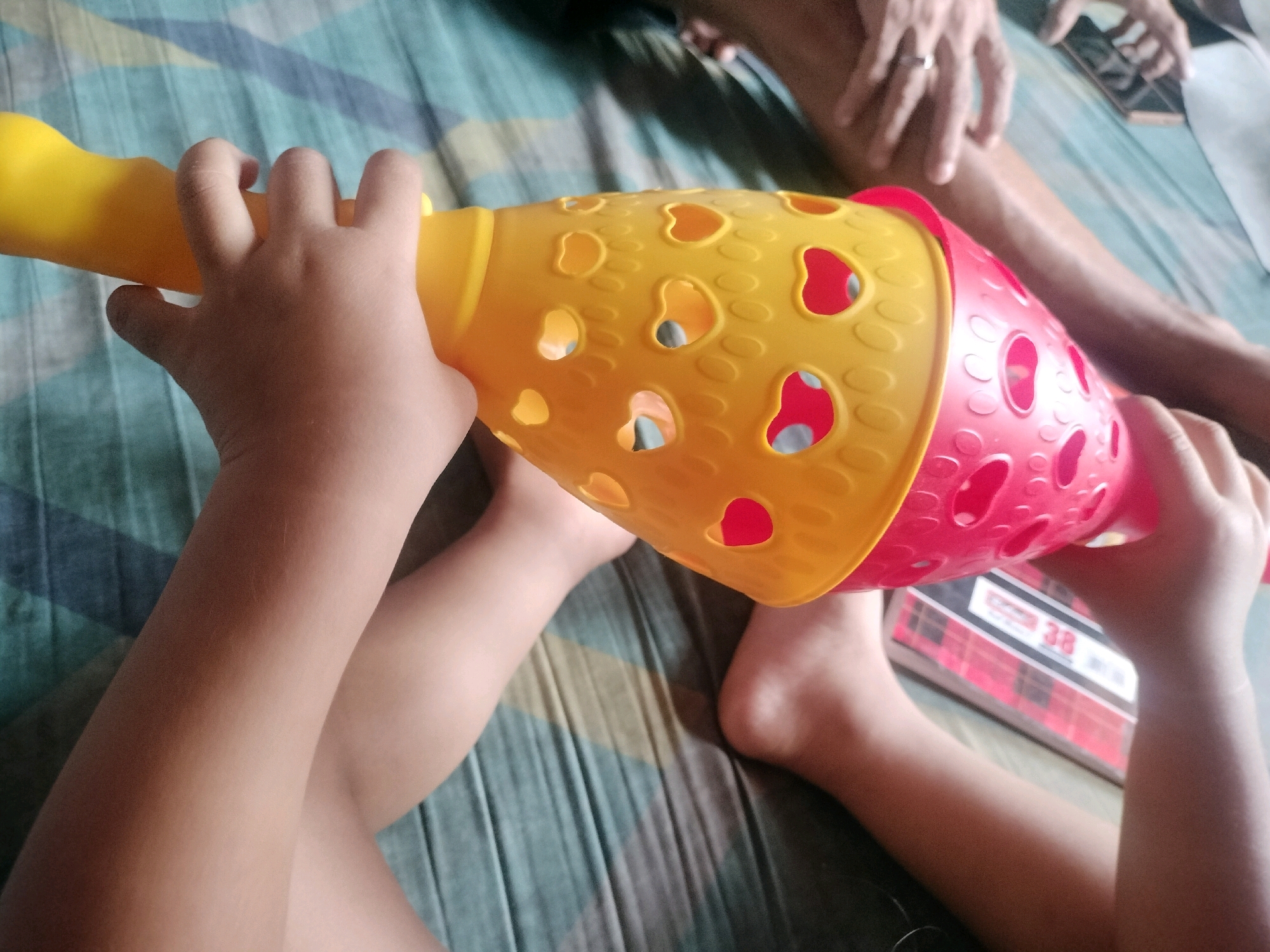In South Jakarta, Indonesia, a child's hands are holding a yellow and red plastic toy, possibly a ball popper or a stacking toy, with heart-shaped cutouts. The toy is positioned horizontally across the frame. The child's legs and feet are visible, suggesting they are lying down. In the background, adult hands can be seen, one wearing a ring, and a smartphone. The scene takes place on a bed with a striped blue and green patterned bedsheet. A book with a red and white checkered cover is partially visible under the toy.