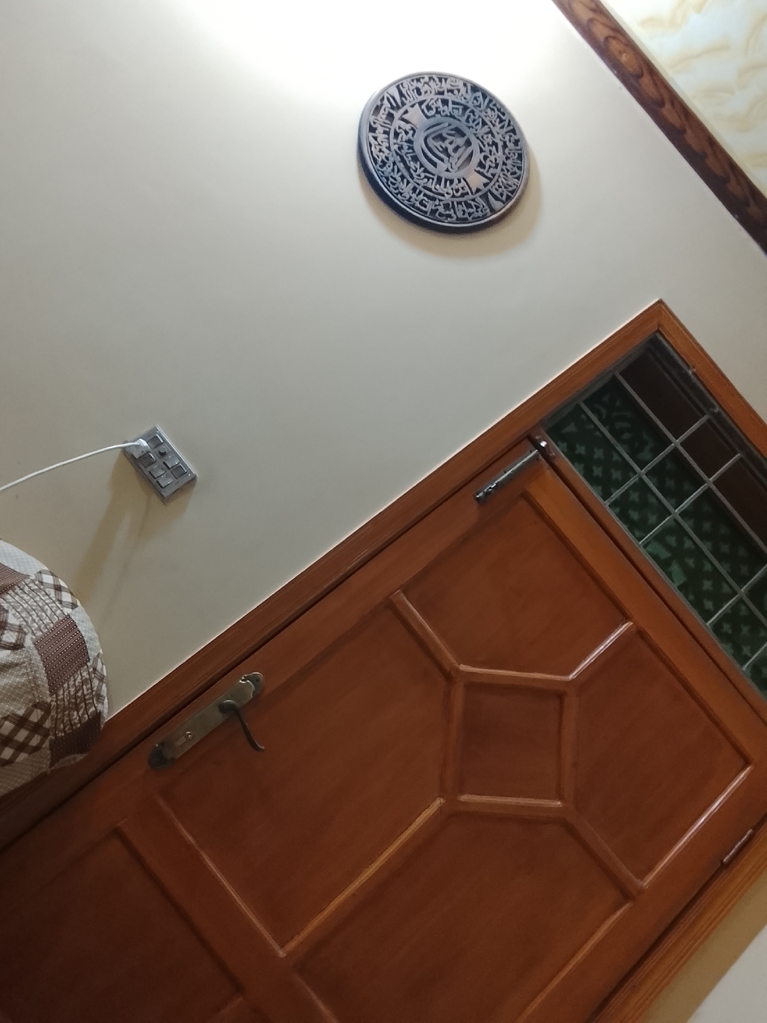 The image shows an interior scene. A brown wooden door with a geometric panel design is positioned diagonally in the foreground. Above the door is a wall with a power outlet and a circular Islamic wall hanging. The image was taken in Multan, Pakistan.