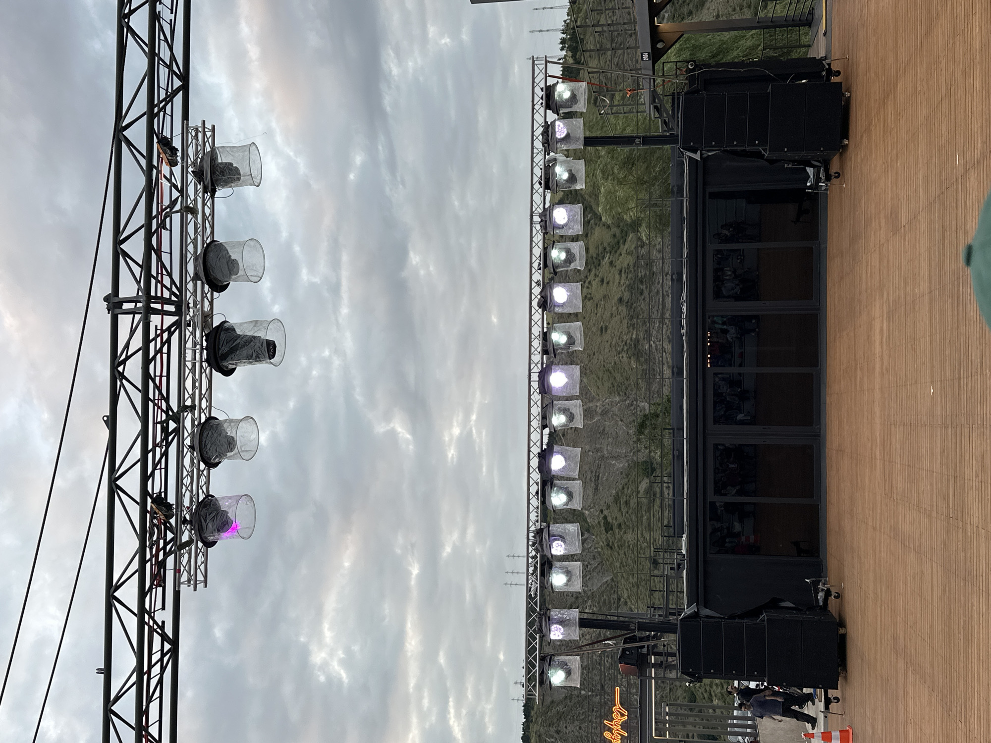 An outdoor stage setup is visible with a wooden floor in the foreground and a cloudy sky in the background. The stage features a black structure with large glass doors, flanked by stacks of black speakers. Overhead, two rows of lighting rigs are suspended from metal trusses. The upper rig holds several large, cylindrical lights covered in clear plastic, with some emitting a pink glow. The lower rig supports multiple smaller stage lights, some of which are illuminated with various colors. To the left of the stage, a neon sign with cursive text is mounted on a metal structure. Several people are present, including two individuals walking on the left side of the image and a group of spectators visible inside the glass-enclosed area of the stage structure. The scene appears to be set in an outdoor amphitheater or event space, with a hillside covered in greenery and trees visible behind the stage. Utility poles and wires are also present in the distance. The overall impression is of a pre-event or setup phase for an outdoor performance.
