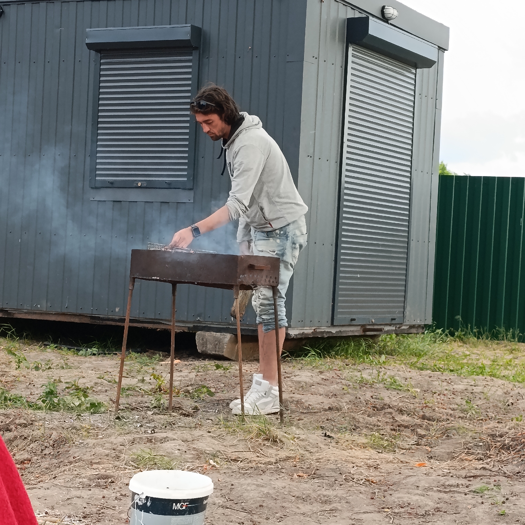 A man with long, dark hair, wearing sunglasses on his head, a light-colored hoodie, and distressed denim jeans, is standing in front of a charcoal grill. Smoke is rising from the grill, and the man's hand is visible, appearing to be attending to something on the grill. He is wearing white high-top sneakers. Behind him is a grey, corrugated metal building with a roll-up door or shutter. To the right of the building is a tall, green corrugated metal fence. The ground is a mix of dirt and dry grass. In the foreground, to the left, part of a red object and a white bucket with black lettering are visible. The scene appears to be outdoors, possibly in a rural or industrial area, within the city of Ladyzhyn, Ukraine.
