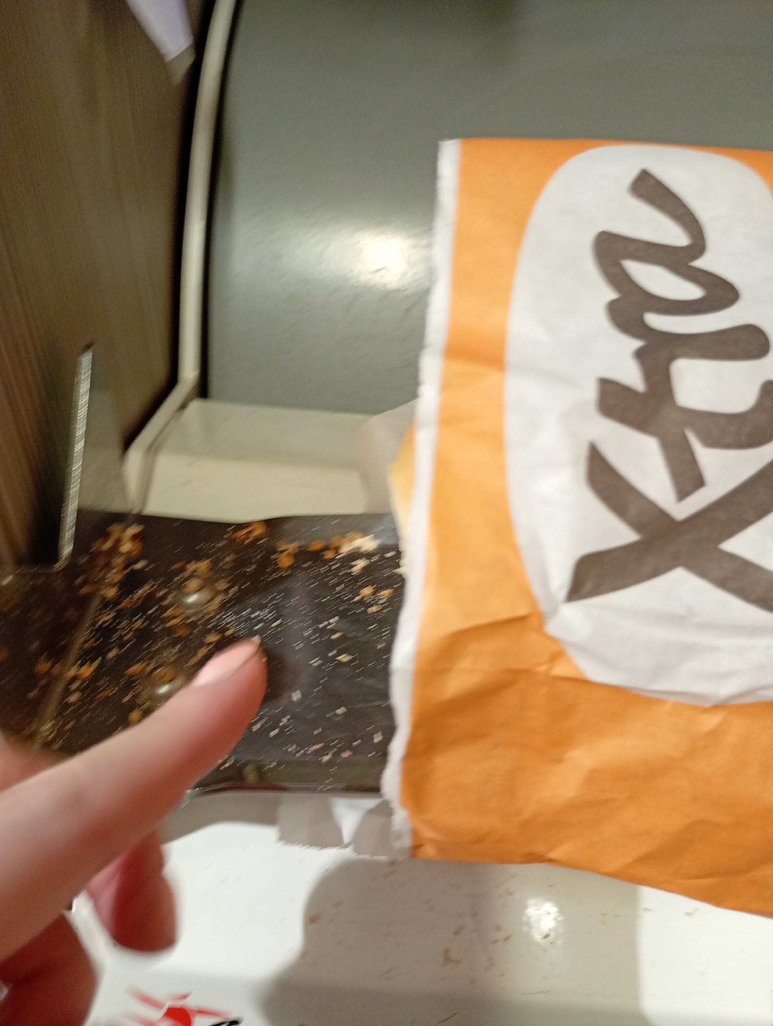 The image shows a close-up view of a finger with light-colored nail polish touching a dark surface. The dark surface appears to be a tray or a part of a machine, with scattered crumbs or small pieces of food on it. To the right of the finger, a crumpled orange and white paper bag with dark lettering is visible. The lettering on the bag is partially obscured but appears to be in a stylized font, possibly indicating a brand or product name. The background is out of focus, showing a blurred vertical surface to the left and a gray surface above. The overall scene suggests a casual setting, possibly a fast-food establishment or a place where food is served. There is no discernible indication of the specific location within a city or any other environmental details.
