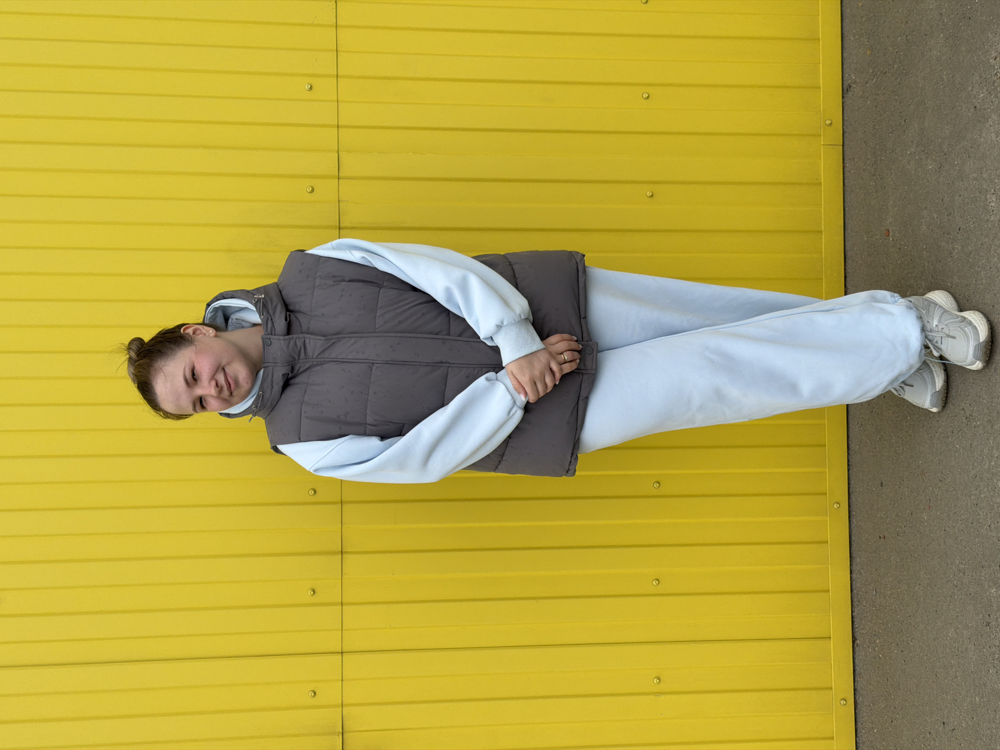 A young woman stands outdoors in Chaikovsky, Russia, captured against a bright yellow corrugated metal wall. The image shows her in a standing pose, smiling gently at the camera with her hands clasped in front of her. She is dressed in casual attire, featuring a dark grey puffer vest worn over a light blue hooded sweatshirt and matching light blue sweatpants. Her feet are in light grey and white sneakers. Her hair is pulled back into a bun. A ring is visible on her left hand. The setting appears to be an urban or industrial outdoor space, with a section of concrete ground visible to the right and beneath her feet. The lighting suggests it is daytime, likely clear and bright. There is no visible text in the image.