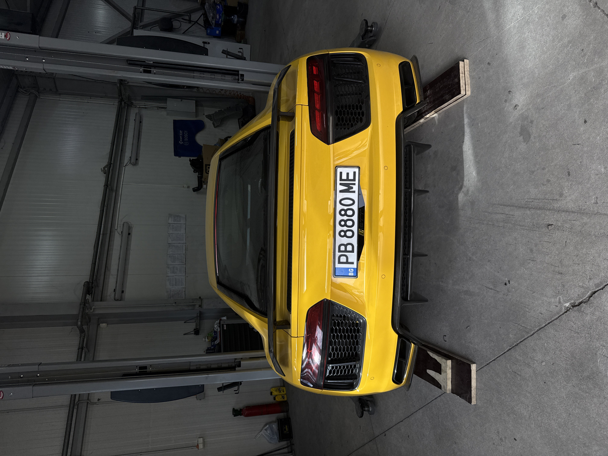 A yellow Audi R8, displaying Bulgarian license plate "PB 8880 ME," is positioned in a garage. The car is elevated on wooden blocks placed under its rear tires. The setting appears to be a car repair or maintenance facility, potentially located in Plovdiv, Bulgaria, given the license plate registration. Garage tools and equipment are visible in the background. The car is shot from behind, highlighting its rear design, including the taillights and diffuser.