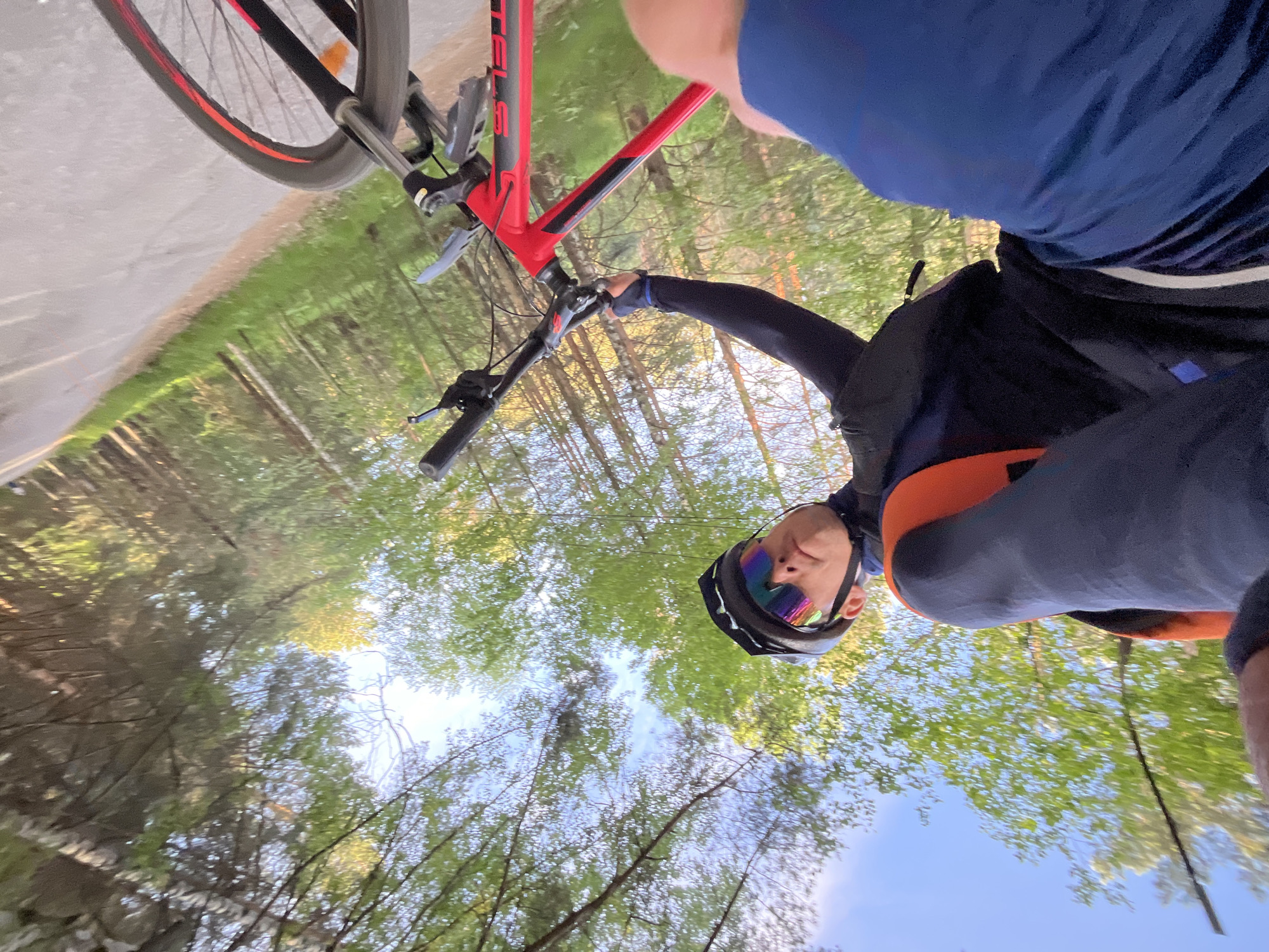 A low-angle view shows a person wearing a cycling helmet and sports glasses, riding a red bicycle on a path surrounded by trees. The person is wearing a dark long-sleeved top and blue shorts. The path appears to be paved, and the trees overhead create a canopy. The image was taken in Chaikovsky, Russia.