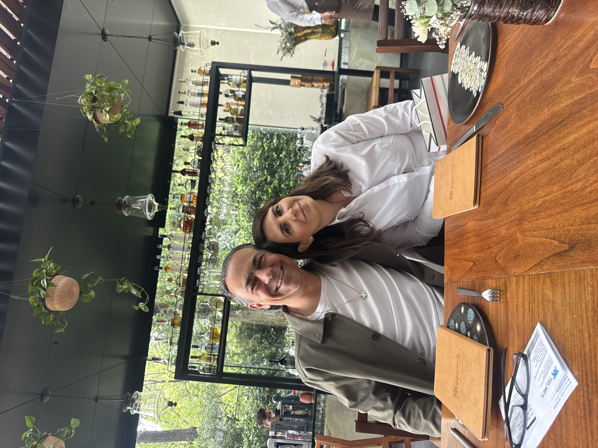 A horizontal, medium shot depicts a man and a woman posing at a wooden table in what appears to be a restaurant or café setting. The location is in Mexico City, Mexico.
The woman is wearing a white, button-down shirt and has dark hair. She is leaning into the man, who is smiling at the camera. The man is wearing a white T-shirt underneath a green jacket, and a silver necklace.
The table is set with plates, silverware, menus, a vase with flowers, and a pamphlet. Behind the couple is a black shelving unit filled with bottles, possibly indicating a bar or beverage display. Outside the shelving unit, there is a view of greenery, suggesting an outdoor or garden setting. Above the couple, hanging plants in pots add to the ambiance.