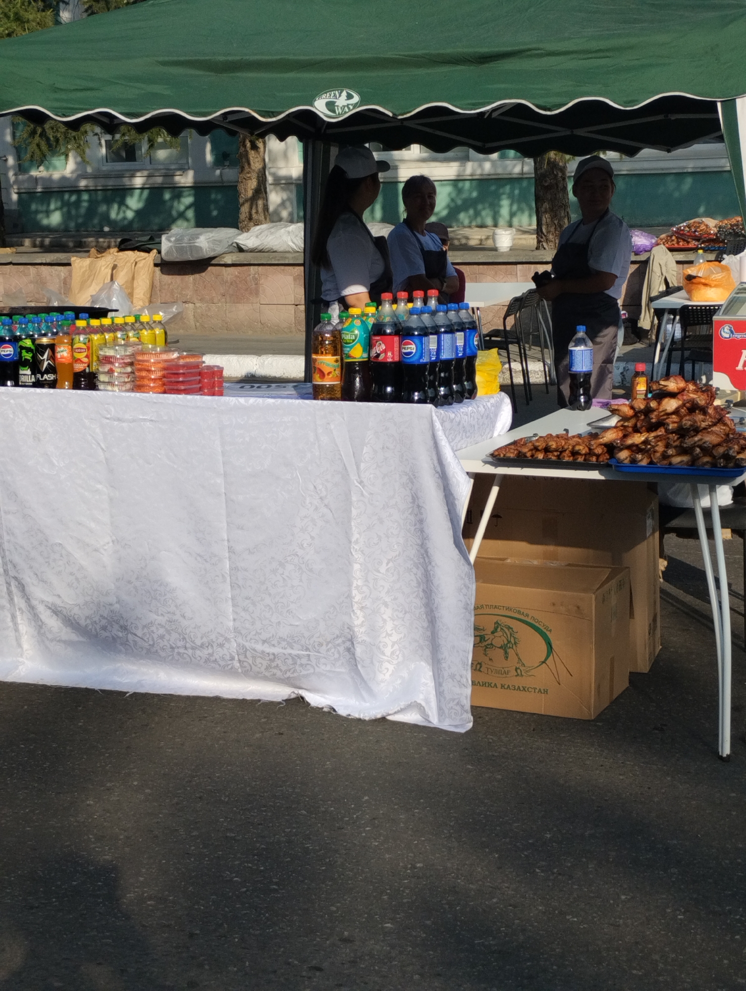 The image depicts an outdoor market stall in Kostanay, Kazakhstan, beneath a green awning. Three individuals, likely vendors, are present. One person, wearing a white cap and blue shirt, stands behind a table laden with various beverages in bottles and plastic containers. To their right, another individual in a white cap and apron is visible. A third person, also in a white cap and apron, is seen further back, holding a bottle and a black object. A table to the right displays a large pile of cooked chicken pieces on platters, with a cardboard box and other items underneath. The ground is asphalt.
