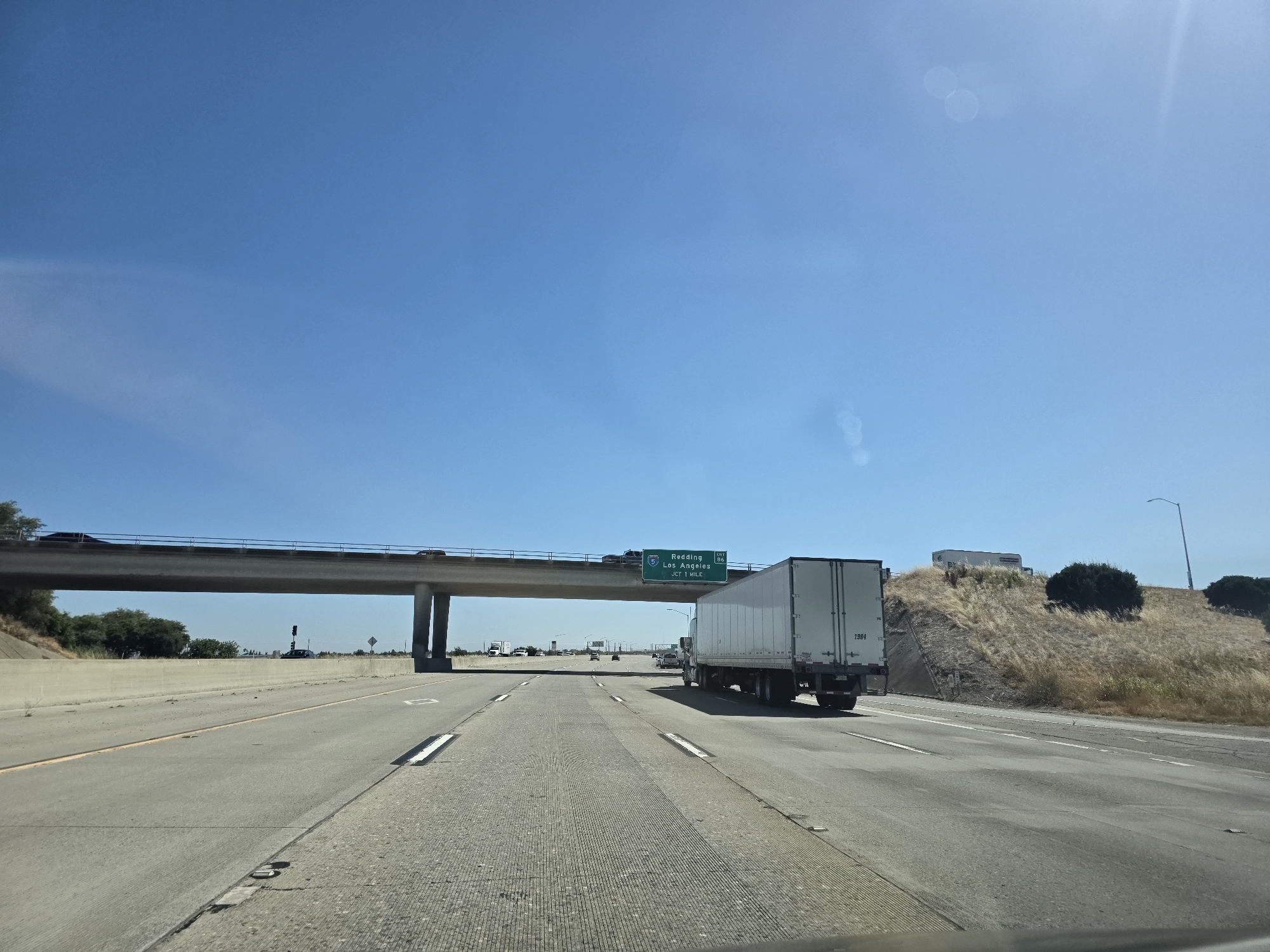 This media file captures a view from inside a vehicle traveling on a highway. The scene depicts a bright, sunny day with a clear blue sky. A wide, multi-lane highway stretches ahead, with concrete barriers lining the sides and intermittent reflective markers on the road.
A prominent element is an overpass with vehicles driving on it, supported by large concrete pillars. To the right of the highway, a dry, grassy hill rises, dotted with sparse bushes. A white semi-truck is in the right lane, moving in the same direction as the viewer's vehicle.
Overhead, a green highway sign is visible. It indicates "Redding" with exit number "5" and "Los Angeles" with exit number "86". Below this, it reads "Jct 99 Mls". This text provides crucial location cues, suggesting the vehicle is on a highway in California, likely heading towards or passing through areas that connect to these cities.
Further down the highway, several other vehicles, including trucks and cars, are visible, indicating moderate highway traffic. The overall impression is a typical day of highway travel in a warm, dry climate, likely California given the signage. The time of day appears to be midday due to the strong sunlight and shadows. No people are clearly visible, reinforcing the perspective from within a vehicle.