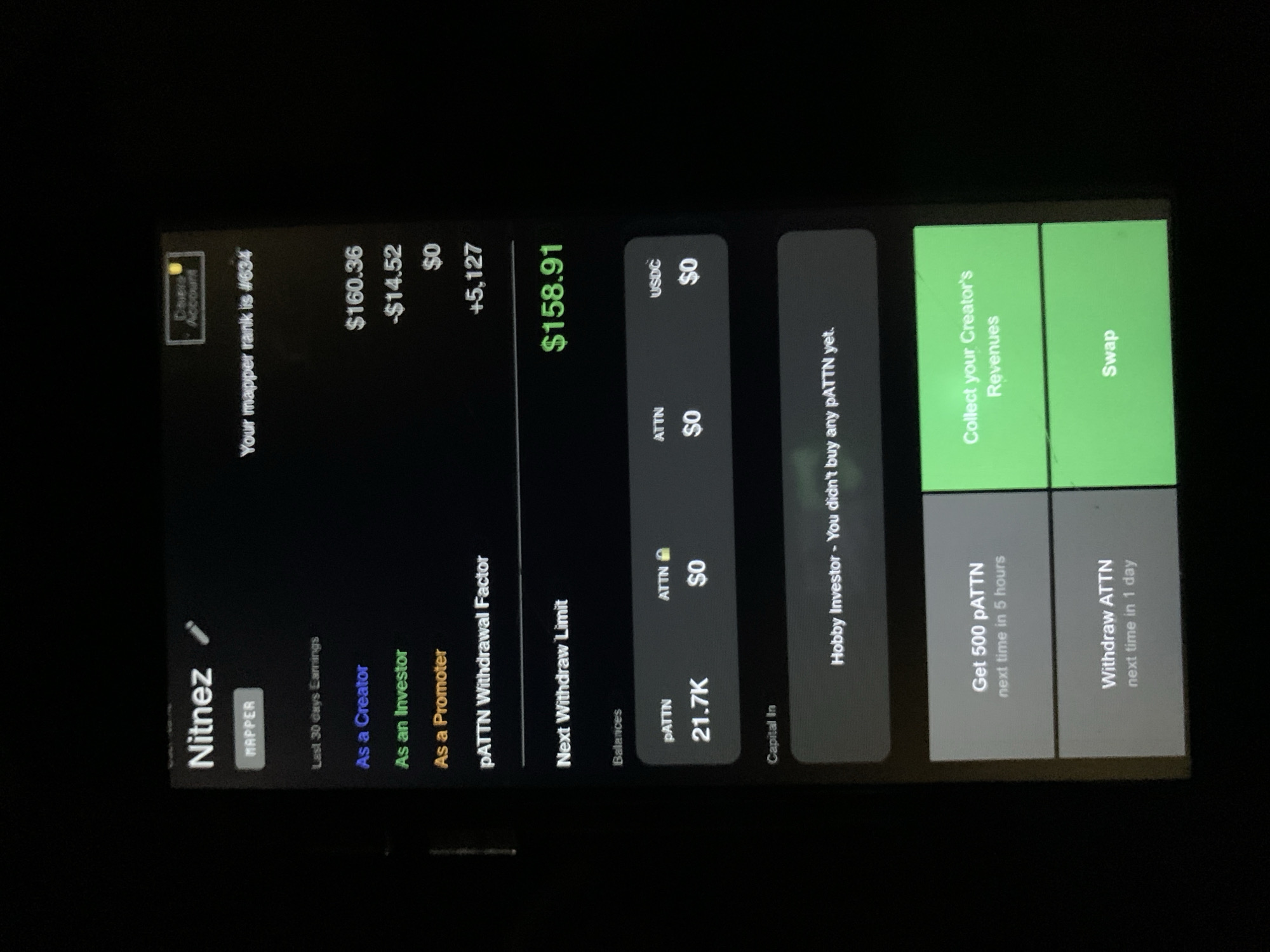 The media file presents a close-up, slightly rotated view of a mobile phone screen displaying a dark-themed application interface. The surrounding environment is dark, suggesting an indoor setting, possibly at night or in a dimly lit room. No individuals are visible.
The application appears to be a personal dashboard for a creator or investor platform. The user's name is "Nitnez," identified as a "MAPPER" with a rank of "#634." The screen details "Last 30 days Earnings," showing "$160.36" as a Creator, "-$14.52" as an Investor, and "$0" as a Promoter. An additional value of "+5,127" is listed, with a net total of "$158.91" highlighted in green.
Under "Balances," the user holds "21.7K pATTN," and "$0" in "ATTN" and "USDC." "Capital In" for "ATTN" is also "$0." A message indicates "Hobby Investor - You didn't buy any pATTN yet." Action buttons include "Collect your Creator's Revenues" and "Swap," both highlighted in green. Further options are "Get 500 pATTN next time in 5 hours" and "Withdraw ATTN next time in 1 day," presented on grayed-out buttons with timers. A "Close Account" button is visible in the top left corner. The scene is set in Rafin Par, Nigeria.