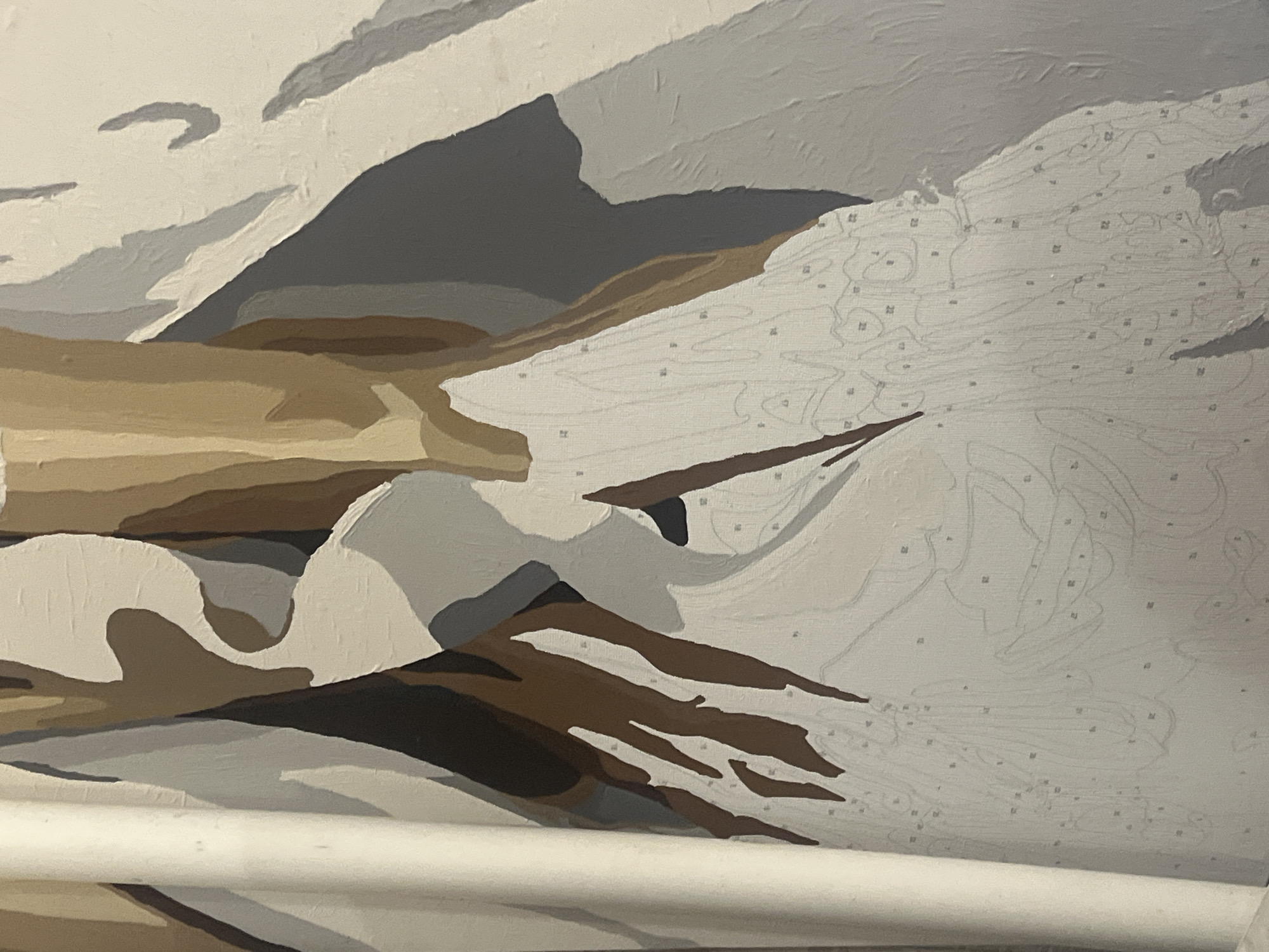 The image shows a close-up of a paint-by-numbers canvas depicting an abstract or semi-abstract subject. The canvas is partially painted with large blocks of color, including white, beige, light gray, dark gray, and brown. Many areas of the canvas are still unpainted and show faint numbered outlines. A vertical white cylindrical object is visible on the left side of the image, possibly a rolled-up canvas or part of a studio setup. There are no people, animals, or recognizable landmarks in the image, and therefore no connection to Saratov, Russia can be determined. The scene is indoors, likely in a studio or hobby space.