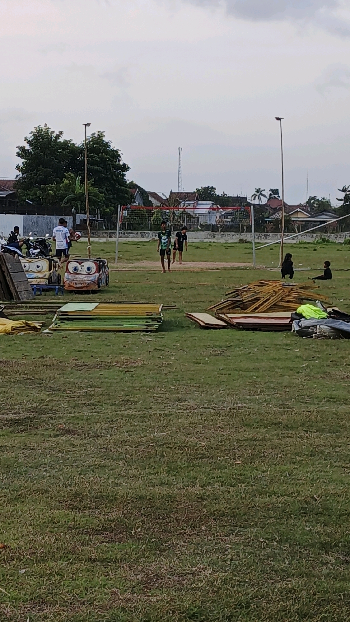 The image depicts an outdoor scene in the city of Boyolangu, Indonesia, likely a public field or park. In the foreground, stacks of colorful, flat objects, possibly wooden planks or panels, are arranged on the grass. Further back, a volleyball net is set up, and several individuals are present. Two young males, wearing athletic attire, are walking on the field near the net. To the left, a person in a white shirt is standing near a motorcycle, and two cartoonish, vehicle-shaped structures, one resembling a car and the other a similar playful design, are situated on the grass. In the background, residential buildings and trees are visible under a cloudy sky. On the right side of the field, two children are sitting on the grass, with one appearing to be looking towards the other figures. A pile of wood is also visible on the right side of the image.