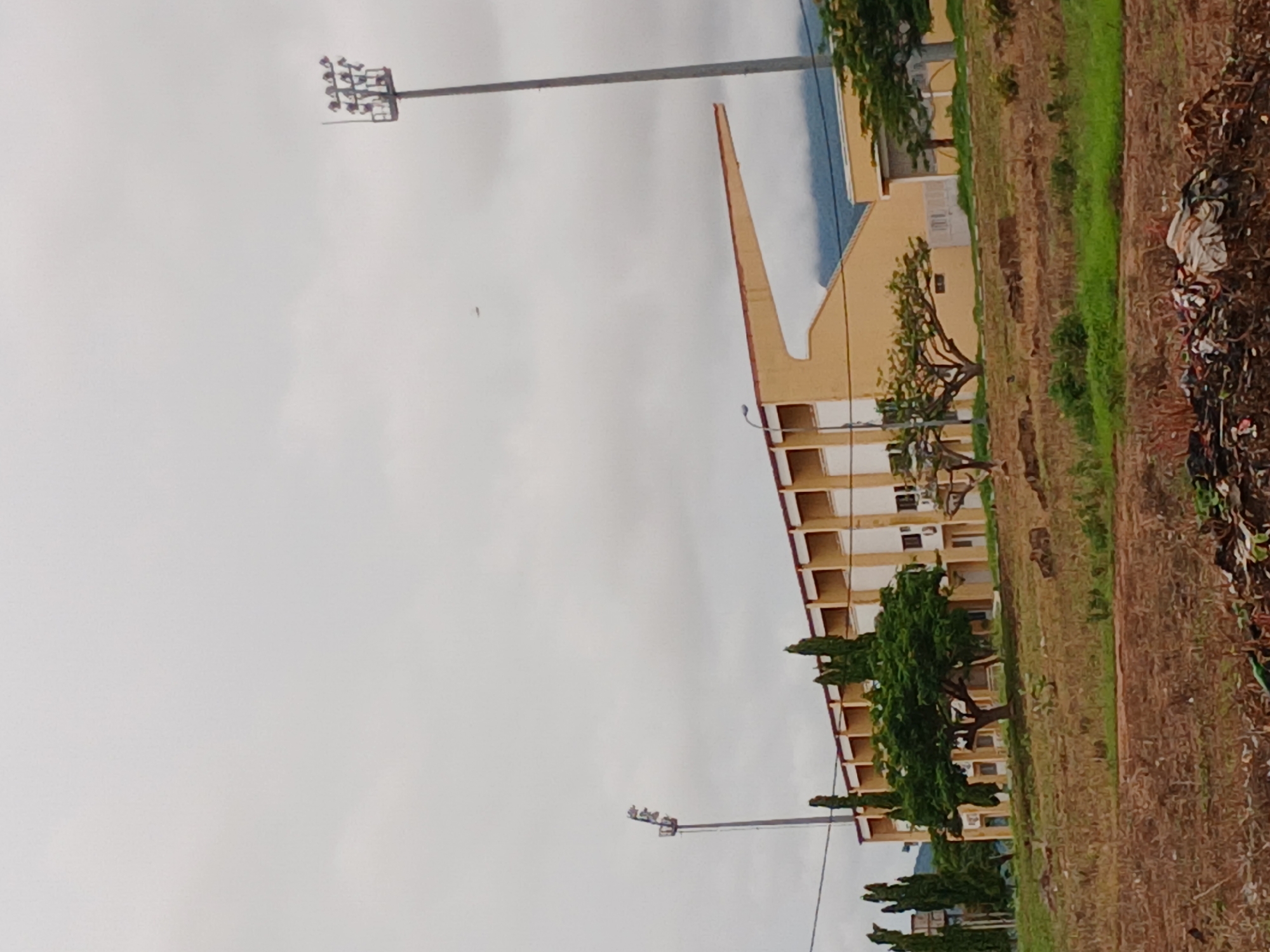 The image depicts a stadium in Howayi, Nigeria, under an overcast sky. The stadium is characterized by its yellow facade and rows of windows. Tall, slender light poles with multiple lamps stand on either side of the structure. In the foreground, a field of dry, brown grass with patches of green hints at a natural, possibly arid, environment. A pile of debris, including plastic bags and other refuse, is visible at the bottom of the frame. Several trees, some tall and slender and others more rounded and leafy, are scattered around the stadium and in the foreground. In the distance, faint mountain ranges are visible through the haze.