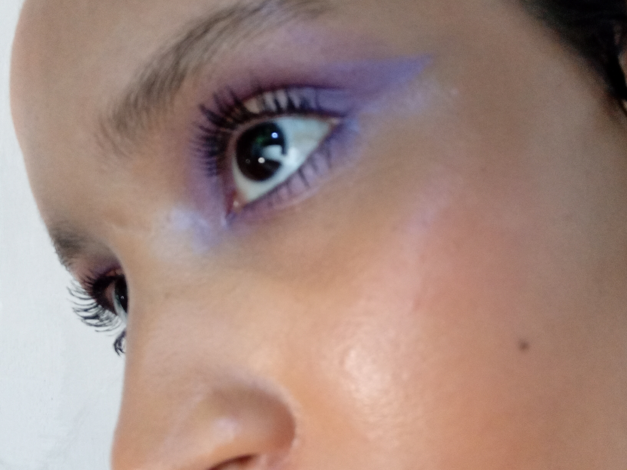 A close-up, angled portrait captures the right side of a person's face, focusing primarily on their eye and surrounding skin. The subject, situated in Parroquia Ocumare, Venezuela, has their eye open, with the pupil reflecting bright light sources.
A vibrant purple eyeshadow is prominently featured, applied across the eyelid and extending outwards in a sharp, winged shape towards the temple. Lighter, possibly iridescent, eyeshadow is visible near the inner corner of the eye and softly blended towards the eyebrow. The eyelashes are long, dark, and voluminous, suggesting the application of mascara or false lashes. The eyebrow above is natural in appearance, with light brown or dark blonde hairs, and appears well-groomed.
The skin on the cheek and around the eye is smooth with a subtle, healthy sheen on the cheekbone, indicating either a natural glow or subtle highlighting. A small, dark mole is visible on the upper part of the cheek, beneath the outer corner of the eye. The overall lighting is bright, casting clear reflections in the eye and illuminating the skin textures. There is no visible text in the image.