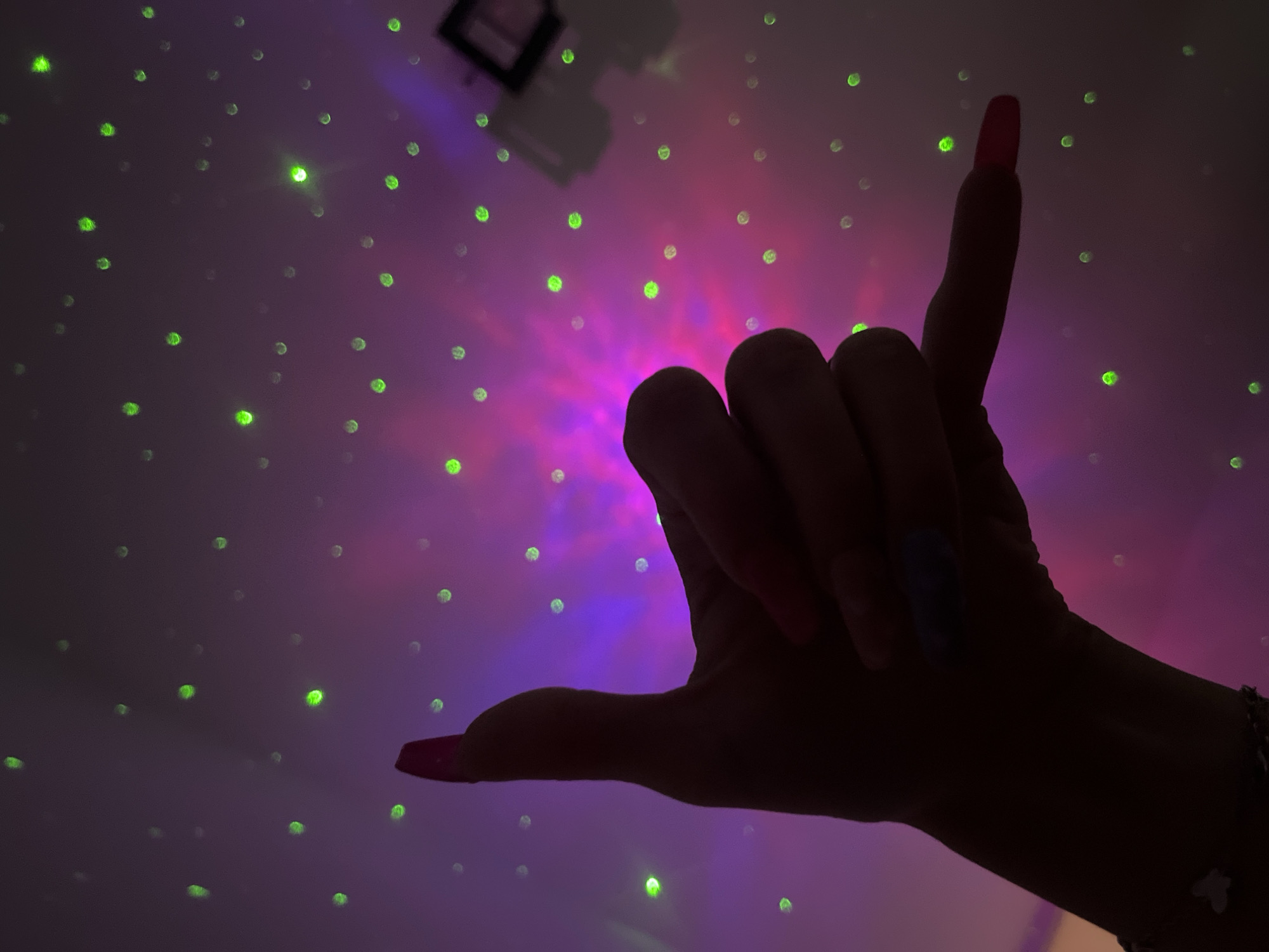 A dark silhouette of a hand with long, pink fingernails is making the "rock on" hand gesture. The background is a purple and pink illuminated surface with small green light dots scattered across it. In the upper-left corner is a dark, rectangular object with two square shapes inside. There are no specific details to suggest the location is Plovdiv, Bulgaria.