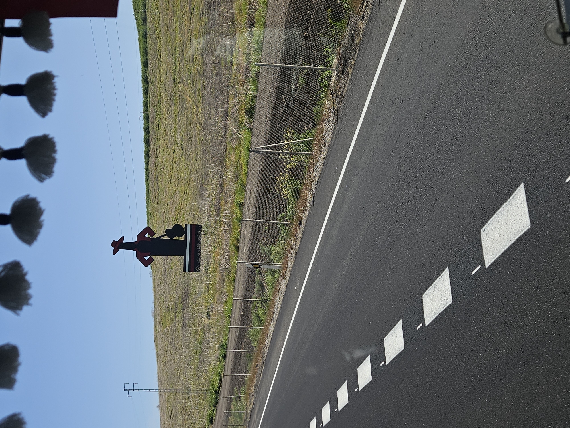 The image shows a road in Los Palacios y Villafranca, Spain. The road is paved with black asphalt and has white dashed lines marking the lanes. A solid white line separates the lane from the shoulder. A chain-link fence runs along the edge of the road, separating it from a dry, grassy field. A sign depicting a silhouette of a person wearing a hat and holding a guitar stands next to the fence. The sky is blue and clear. The foreground includes blurred objects, possibly car decorations hanging near the windshield. Power lines are visible running alongside the field and road.