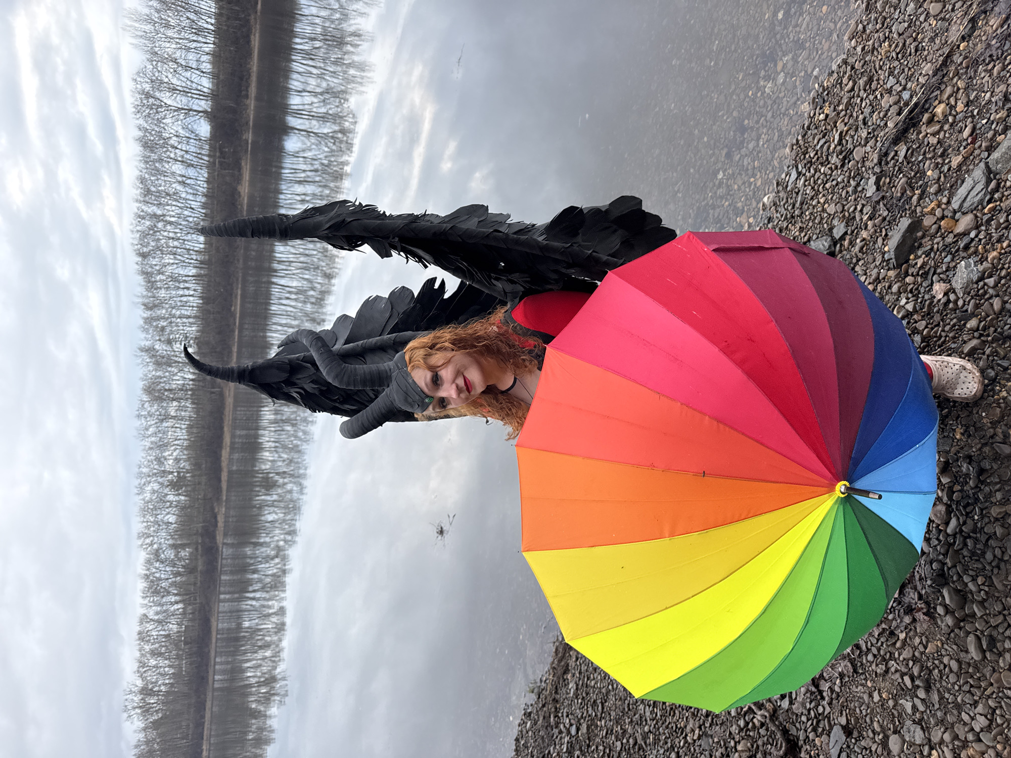 A woman with auburn hair is standing on a gravel shore next to a body of water, possibly the Kama River, in Chaikovsky, Russia. She is wearing black horns and black feathered wings, and holding a closed, rainbow-colored umbrella. The background includes a reflection of trees on the water's surface under a cloudy sky. The shoreline is covered in small pebbles and rocks.