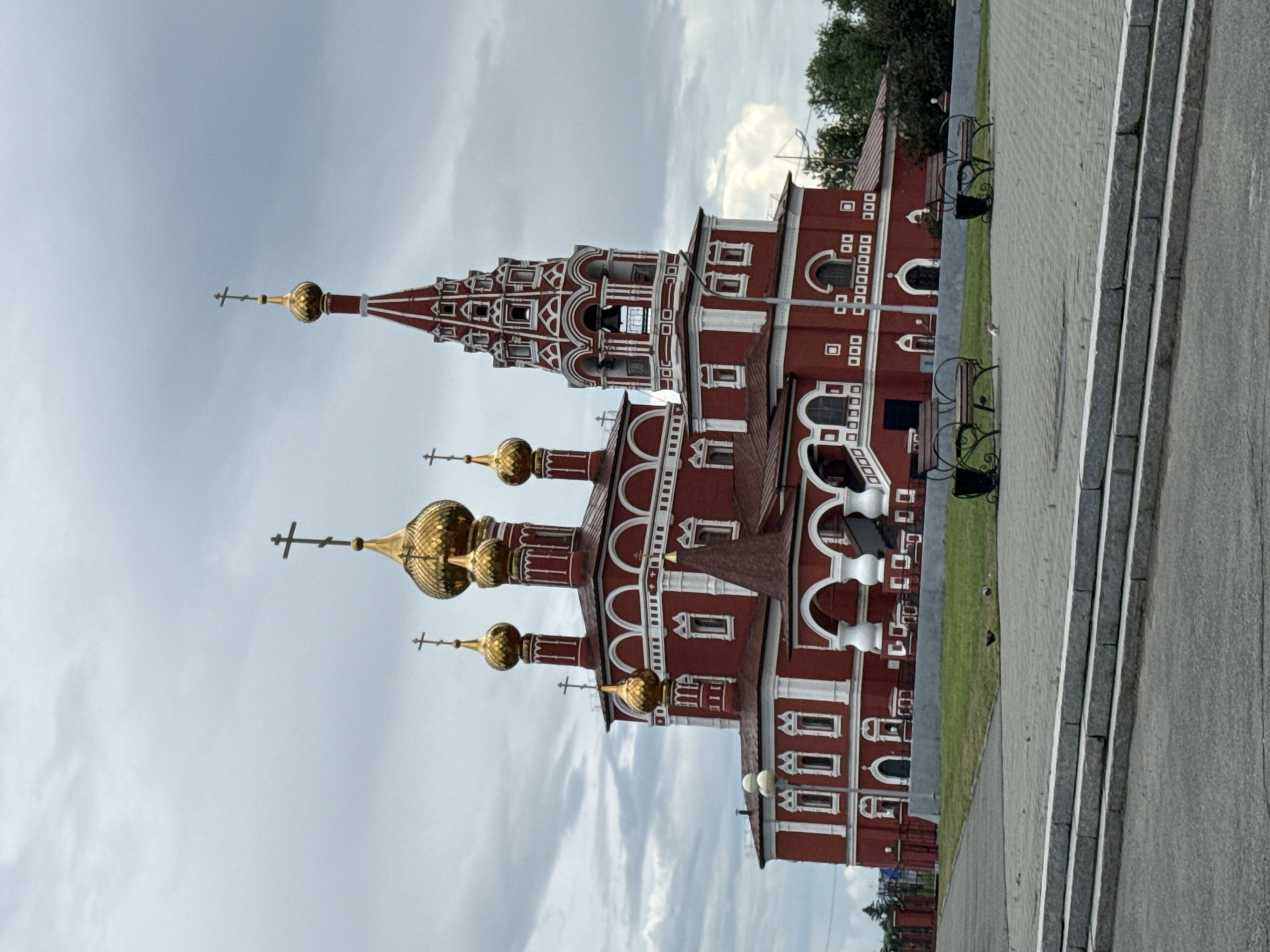 The image shows the Church of the Transfiguration in the city of Kurgan, Russia, under a cloudy sky. The red church features multiple golden domes and a tall bell tower with red and white accents. In the foreground, there are paved walkways, a grassy area with a few benches, and a set of stone steps. The scene is devoid of people.
