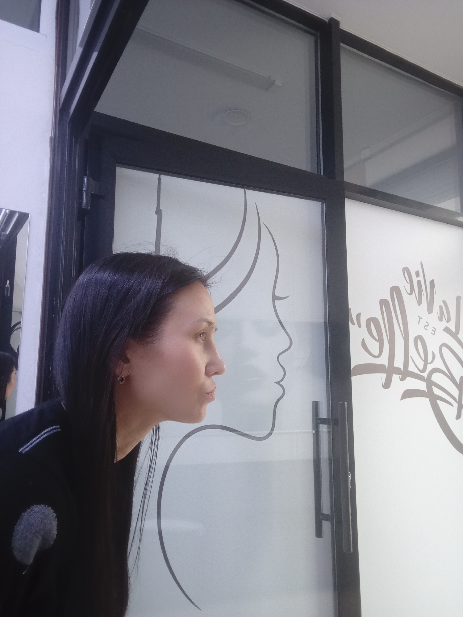 The image captures a woman in profile, positioned next to a modern glass partition or door, likely within an indoor commercial establishment in Izhevsk, Russia. The woman, with long, dark hair, is looking intently to her right (viewer's left), displaying a focused or contemplative expression. She wears a dark long-sleeved top featuring subtle gray or white striped detailing on the shoulder and a soft, circular gray patch on the upper left sleeve. A small, dark earring is visible on her left ear.
The partition consists of dark-framed glass panels, with the lower sections frosted. A prominent feature is a stylized line drawing of a woman's profile etched or applied to the left frosted panel, which aligns closely with the real woman's profile, creating a visually engaging juxtaposition. On the right frosted panel, stylized cursive text is visible. As seen from this side, the text is mirrored, appearing as "эil" (reversed 'Life') and below it, "T2Э" (reversed 'EST') with additional symbols. When read correctly from the opposite side, it would spell "Life" (or a similar word) followed by "EST" and other characters. Dark, horizontal door handles are attached to this panel.
Through the clear upper sections of the partition, the ceiling and interior lighting fixtures are visible, suggesting an indoor setting. To the far left, a reflection of the woman's back and side is seen in an adjacent mirror. The overall scene suggests a clean, contemporary environment, possibly a salon, spa, or office, with the woman seemingly interacting with the decorative elements of the glass partition. The lighting is artificial and consistent with an indoor space.