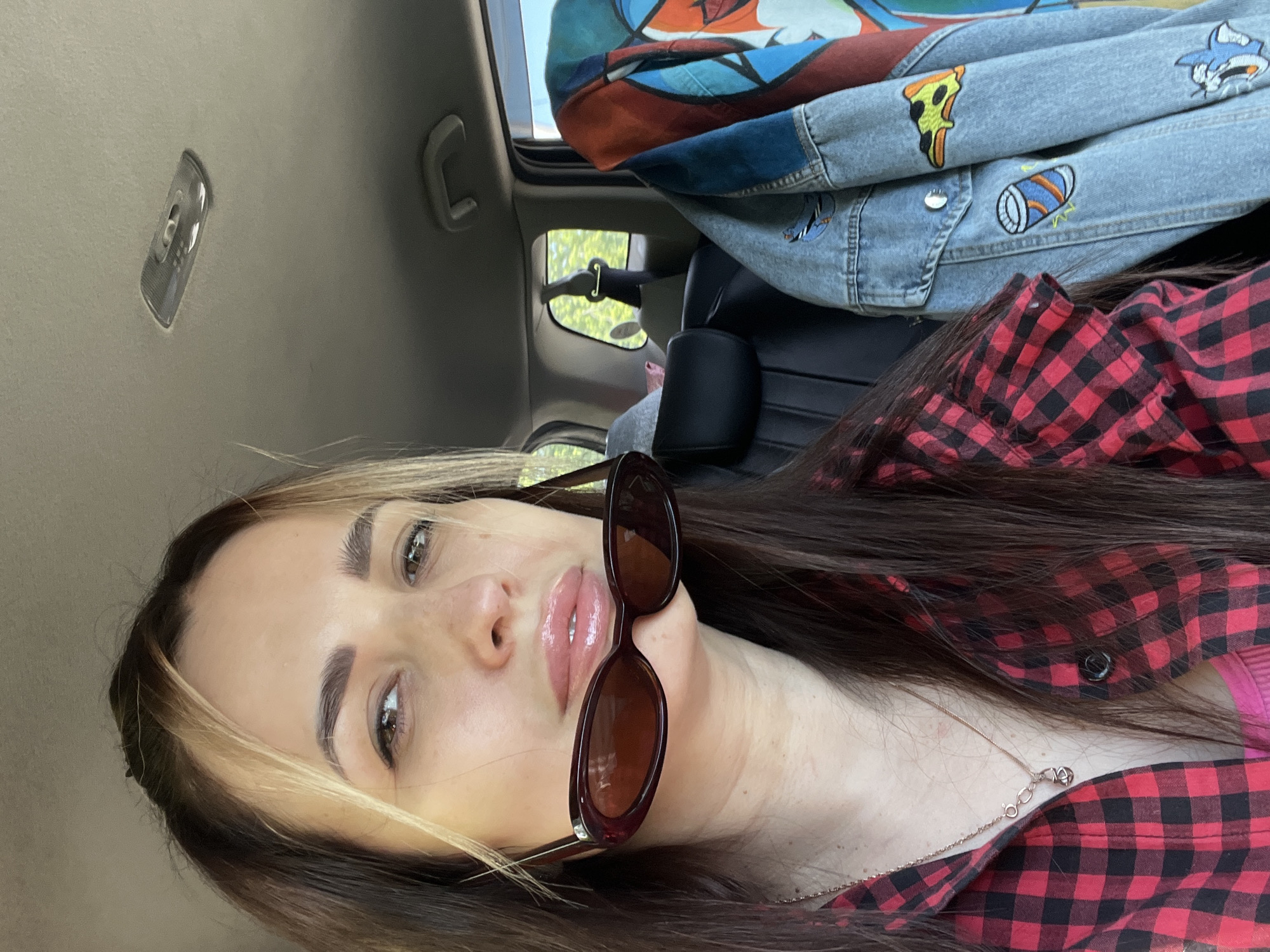 The media file captures a close-up, eye-level shot of a young woman inside a car. The image is oriented vertically, with the woman's head tilted. She has long, dark brown hair with prominent lighter, possibly blonde or caramel, streaks framing her face. Her eyebrows are well-defined, and she has light-colored eyes, appearing greenish-brown, looking directly at the viewer. Her lips are glossy and slightly parted. She wears dark sunglasses perched unconventionally on her nose, resting slightly over her lips.
She is dressed in a red and black plaid button-up shirt, partially open to reveal a hint of another garment underneath, possibly pink. A delicate, thin metallic necklace is visible around her neck.
The setting is the interior of a car. Above and behind her, the car's beige headliner is visible, along with an overhead light fixture and a grab handle. In the background, to the upper right, a vibrant, multi-colored denim jacket with patches (including a pizza slice, a cartoon shark, and a soda can) is folded over a car seat or headrest. Through a car window, bright green foliage from outside is visible, suggesting a natural or suburban environment during daytime. The light appears bright, indicating clear weather. No visible text is present in the image.