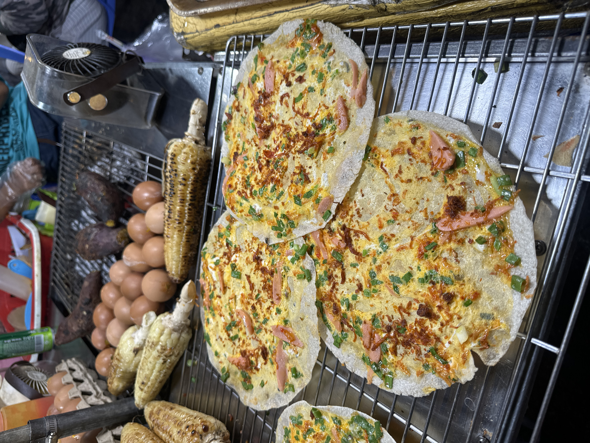 Captured at night, the image depicts a street food vendor in Da Lat, Vietnam. A silver metal rack dominates the foreground, laden with four large, circular rice paper pizzas topped with egg, sausage slices, and herbs. To the left of the pizzas, a selection of cooked corn on the cob, raw brown eggs, and two sweet potatoes are visible on a lower rack. A small, portable fan is clamped to the metal rack. The background is cluttered with hints of other vendors and passersby, creating a sense of a bustling street market.