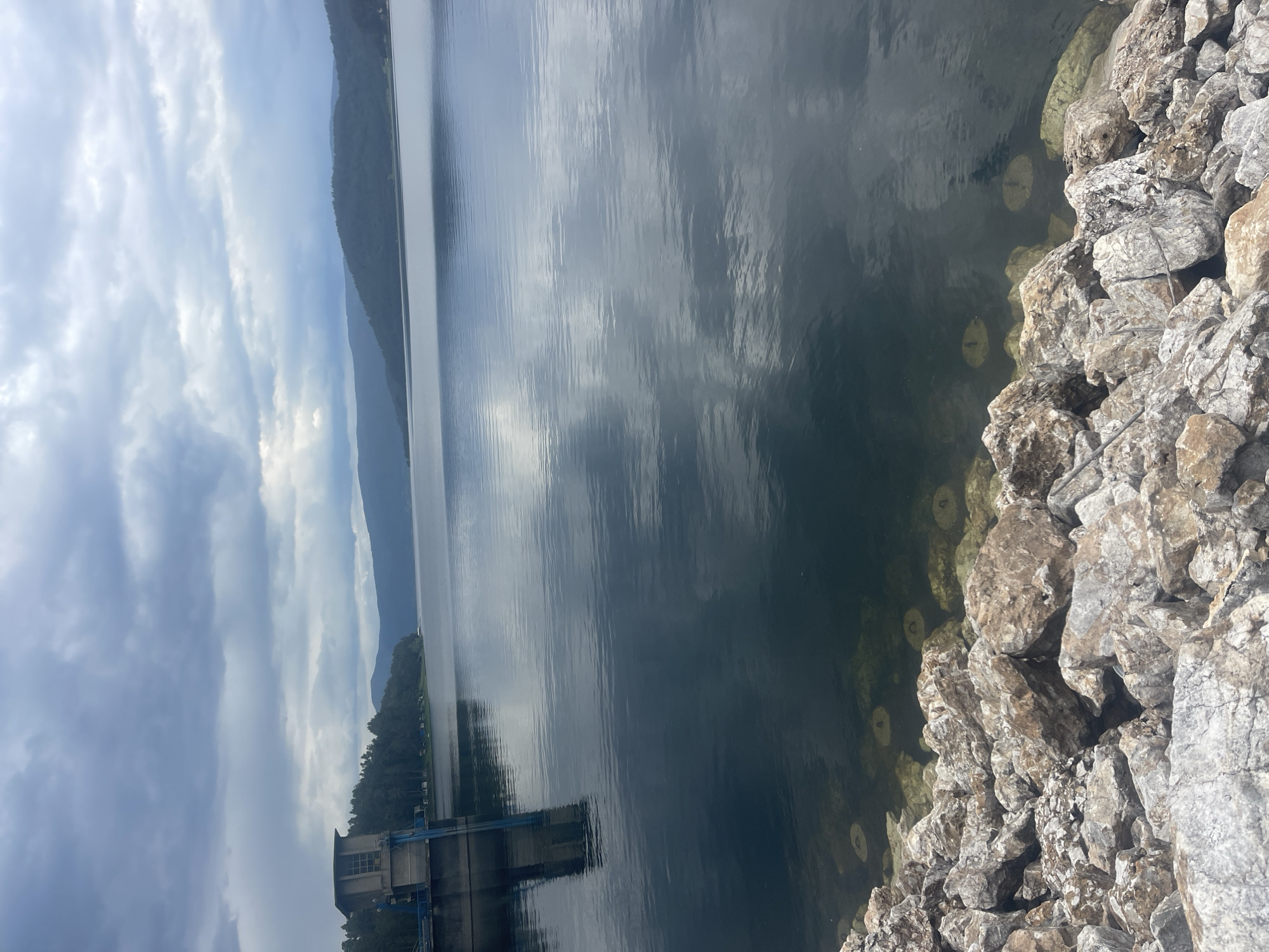 The image shows a body of water next to a rocky shoreline, with a building structure in the distance and a mountain range in the background under a cloudy sky. The location is likely near Sv. Konstantin, Bulgaria. The water appears dark with reflections of the sky and distant mountains. Rocks are clustered along the edge of the water. The building in the distance is a concrete structure.