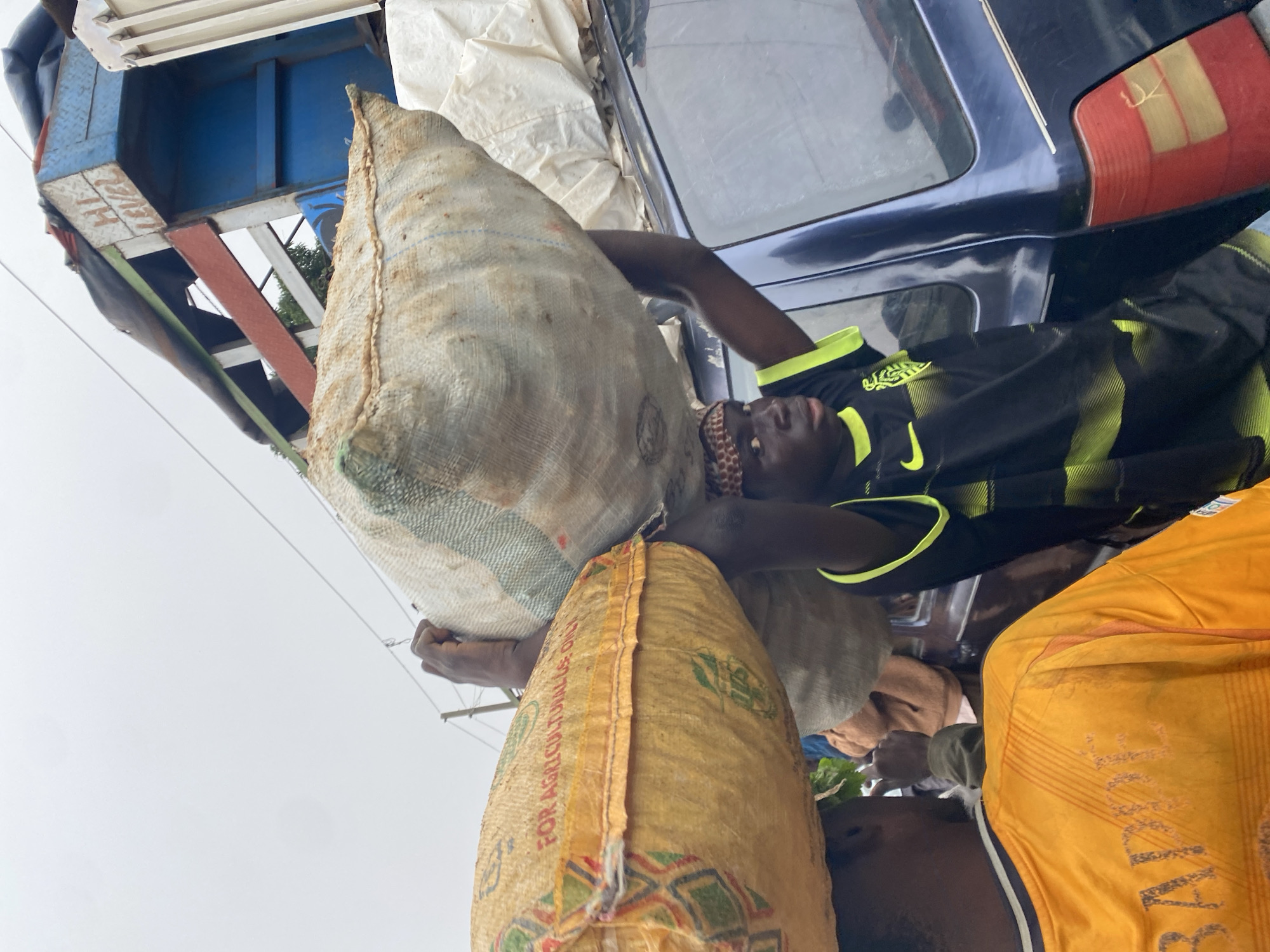 In Katon Rikkos, Nigeria, two young men are seen carrying large sacks of produce on their heads. The man in the foreground is wearing a black and yellow jersey and a patterned headscarf, while the man to his left is wearing an orange jersey with the name "MBAPPE" on the back. They are positioned between a blue truck and a blue vehicle. The truck is laden with more sacks, and its side has the words "HI LEVEL" painted on it. The overall scene suggests a bustling market or transportation hub where goods are being moved. The sky is overcast, indicating potentially challenging weather conditions.