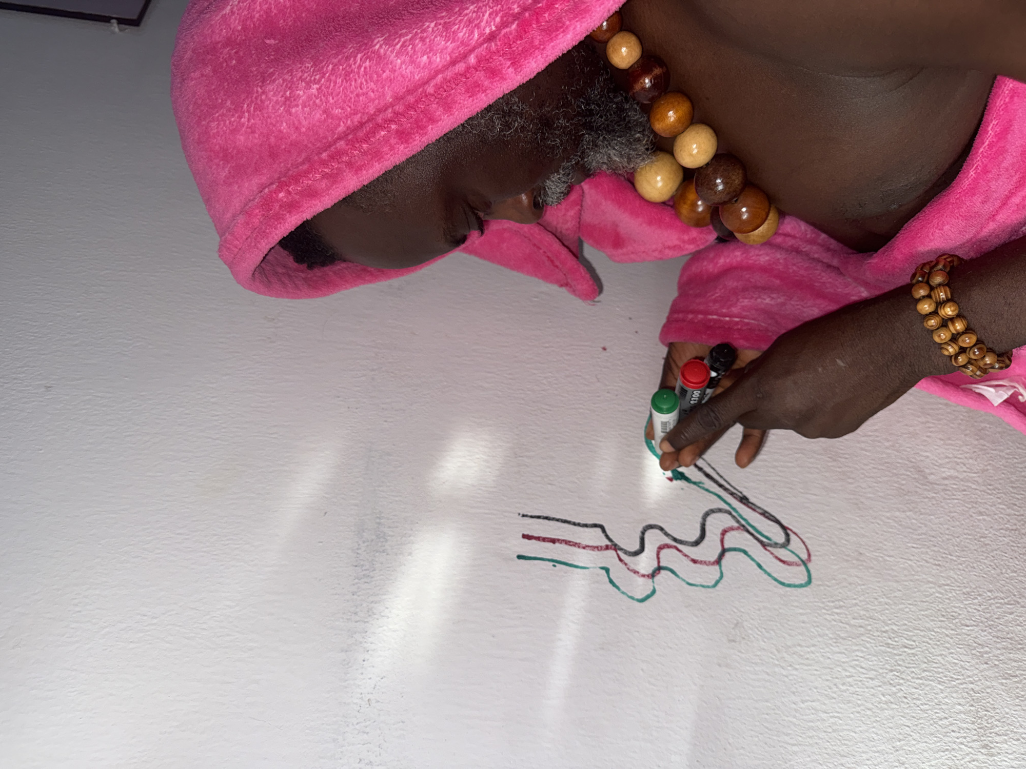 A person wearing a pink hooded robe and wooden bead jewelry is drawing wavy lines on a white wall with green, red, and black markers. The setting appears to be indoors, within a residence. There is no indication of the location being Ado, Nigeria.