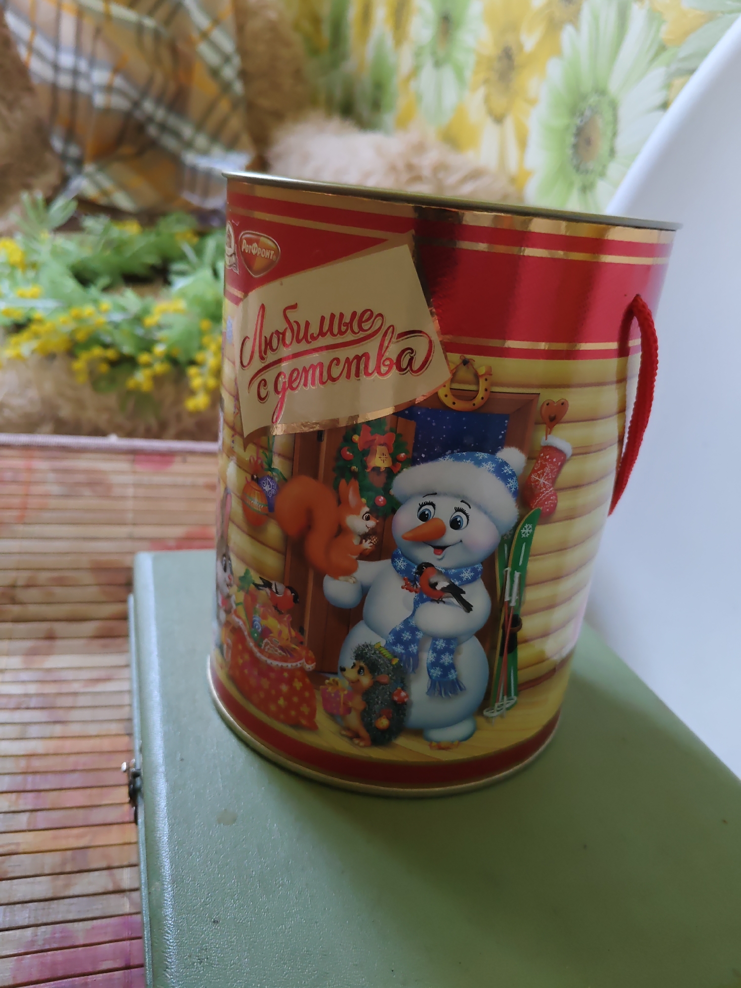 The image displays a cylindrical tin with a Christmas-themed design, featuring a snowman, a squirrel, a hedgehog, and a bird. The tin is decorated with red and gold stripes, and has a red string handle. The text on the tin reads "Любимые с детства," which translates to "Favorites from childhood." The tin is placed on a light green book, which in turn rests on a wooden surface. In the background, a furry, beige object is visible, possibly a stuffed animal, and a curtain with a floral pattern. The lighting suggests an indoor setting, likely in a home or a shop, given the decorative nature of the objects. There is no indication that this scene is located in the city of Rostov-on-Don, Russia.