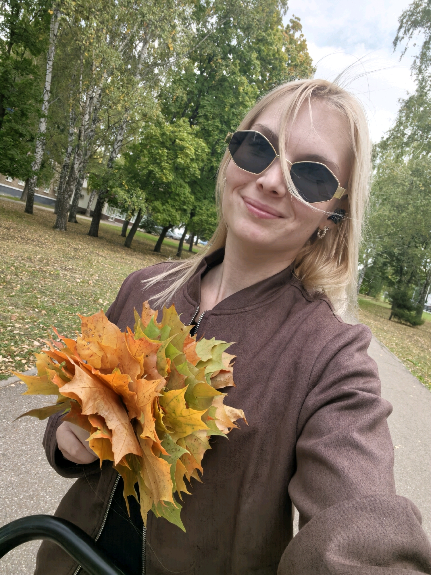 A blonde woman with sunglasses holds a bouquet of autumn leaves in a park. She is wearing a brown jacket. The background shows trees and a pathway. The scene is in Sterlitamak, Russia.