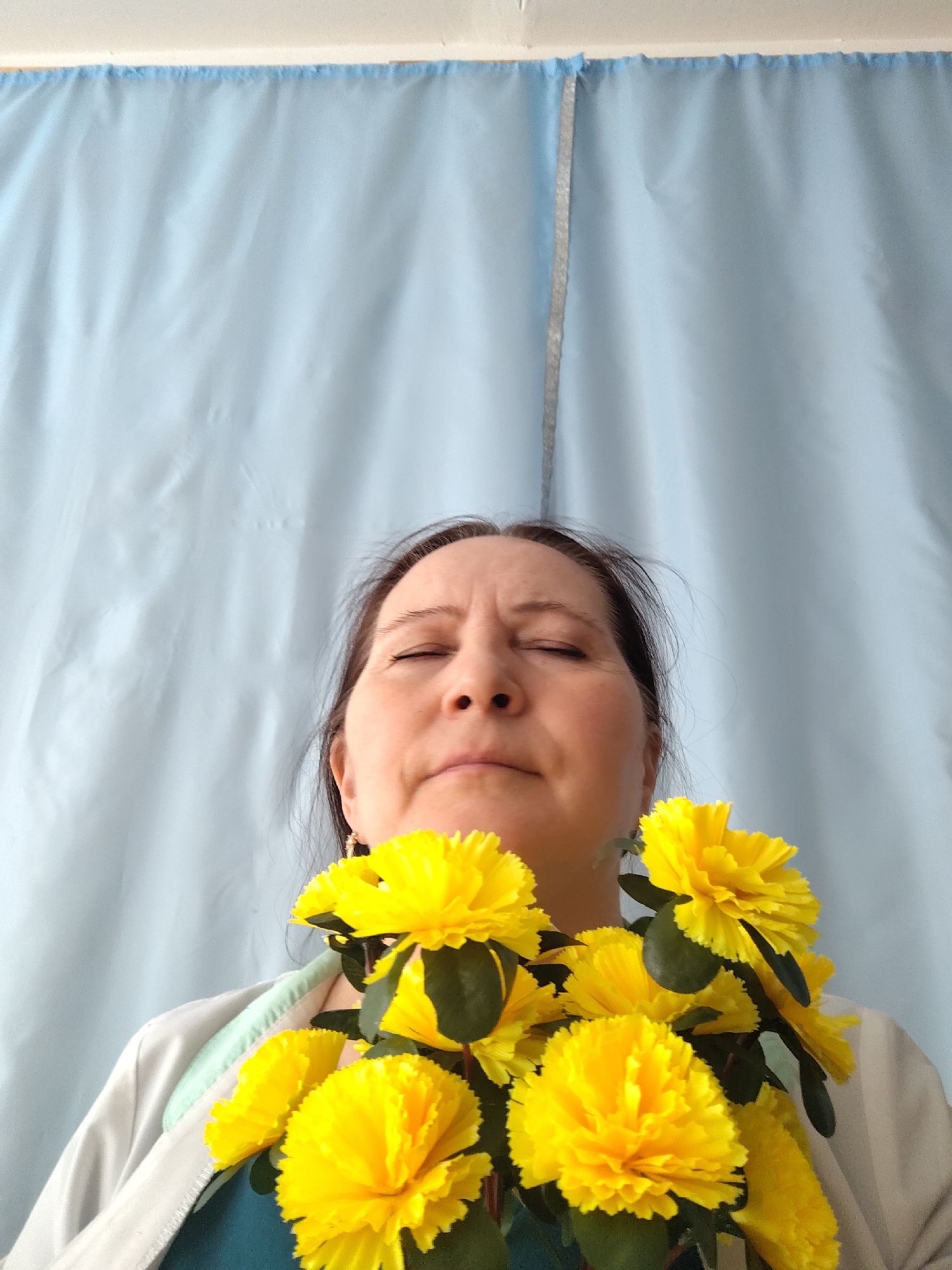 A woman with her eyes closed is standing indoors, holding a bouquet of yellow flowers in front of her chest. She has dark hair pulled back from her face and is wearing a white jacket over a blue shirt. The background is a light blue curtain hanging behind her. There is no indication of the location being Sarapul, Russia within the provided image.